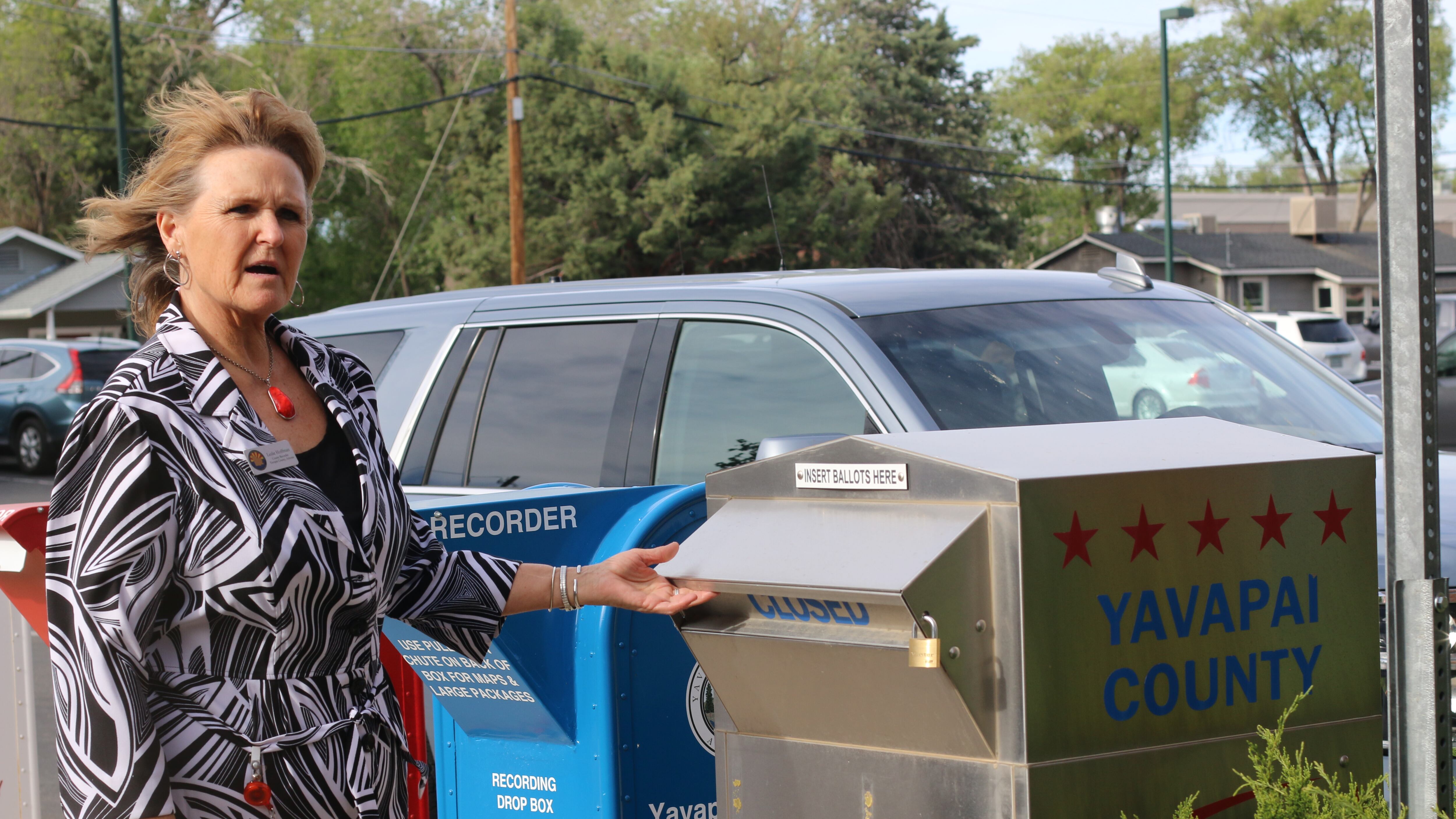 Woman in striped shirt reaches toward a ballot drop box on a sidewalk.
