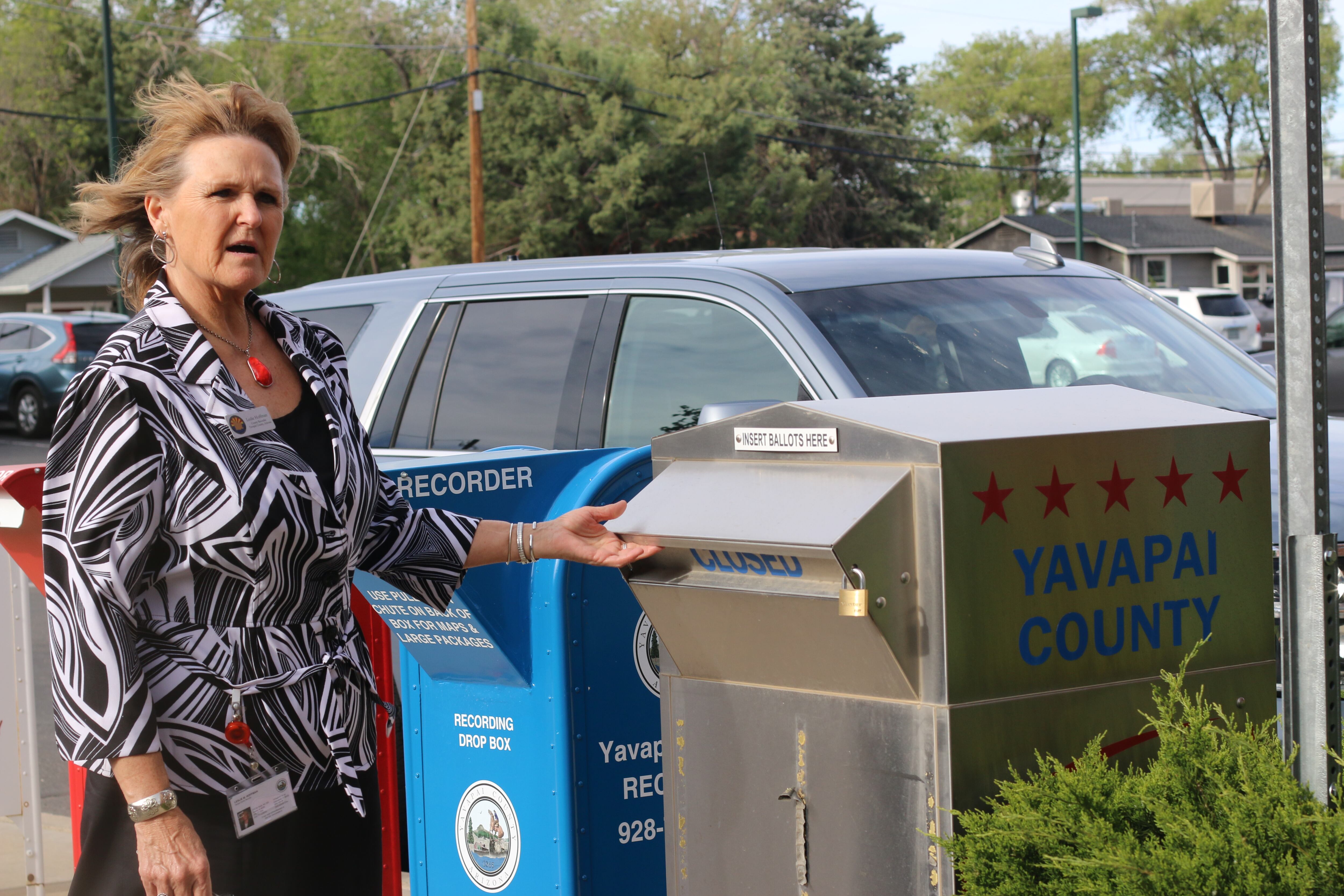 Woman in striped shirt reaches toward a ballot drop box on a sidewalk.