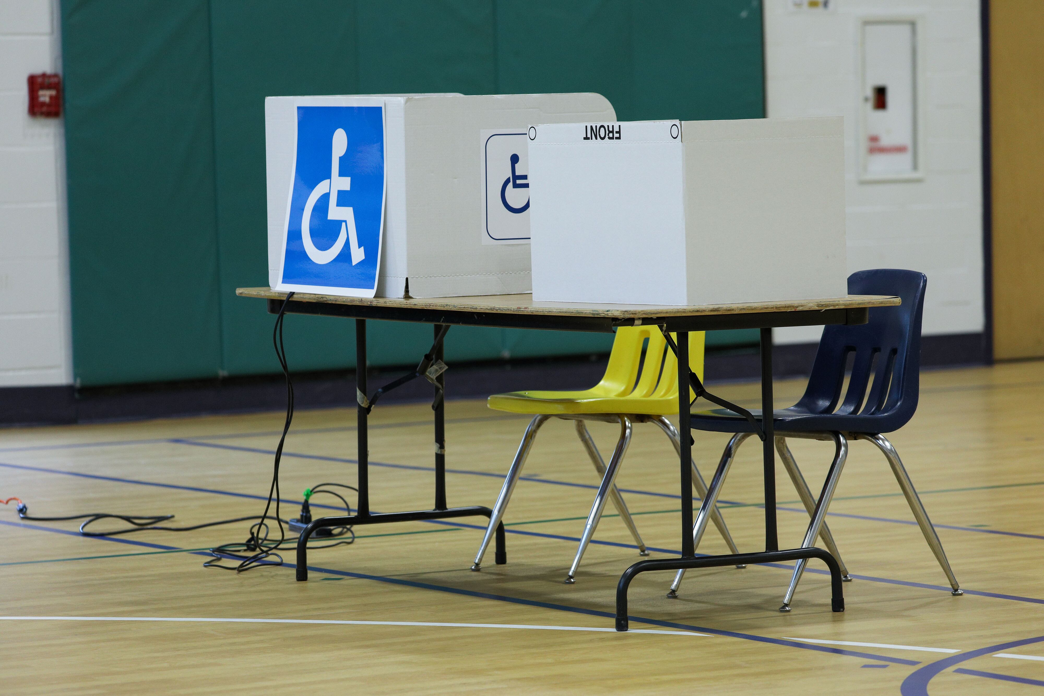 A blue and white handicap sign is on the side of a tan privacy box for voting at a wooden table in the middle of a gym.
