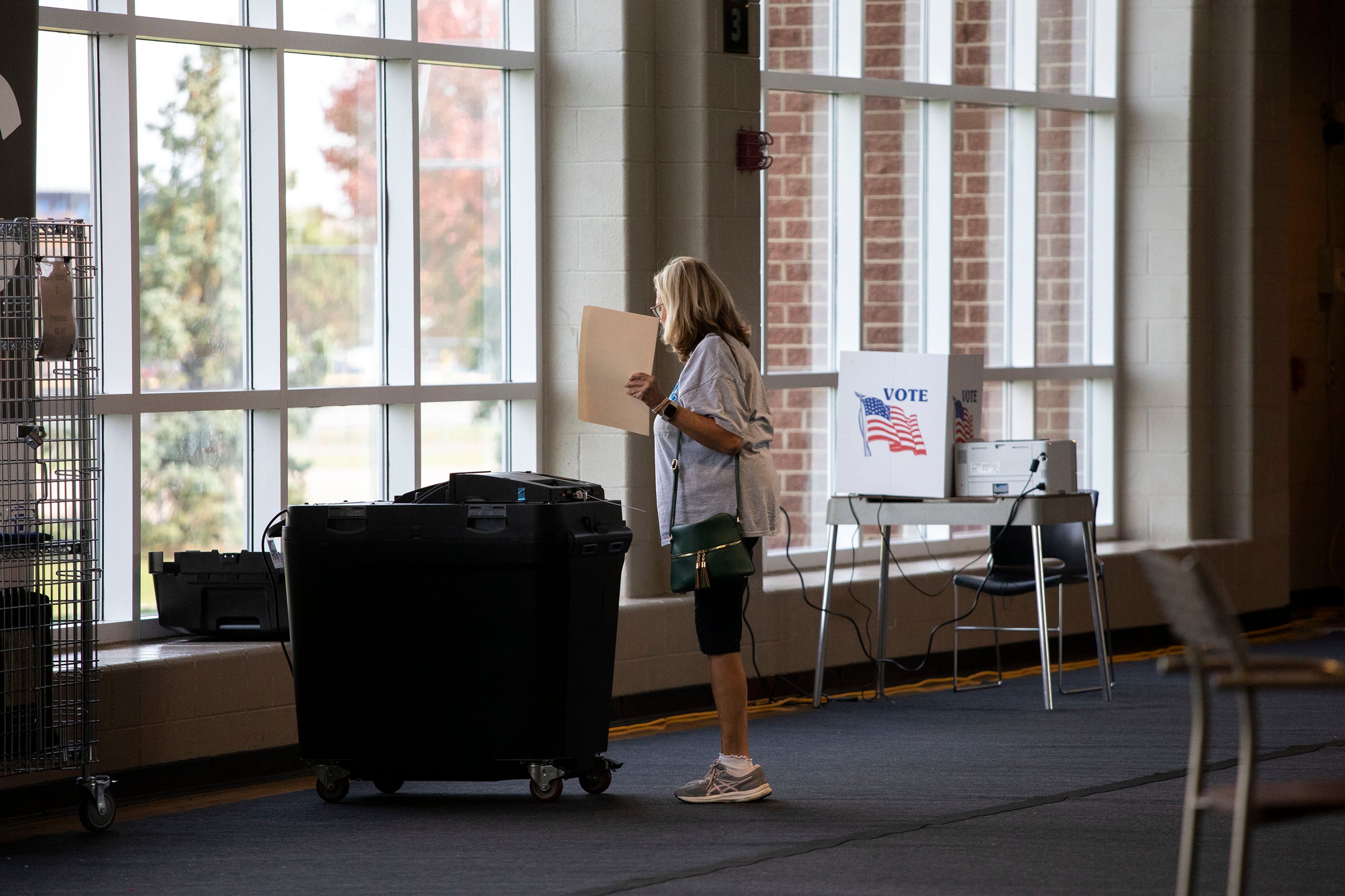 A woman stands in the middle of the frame holding a ballot and standing next to a large black voting machine.