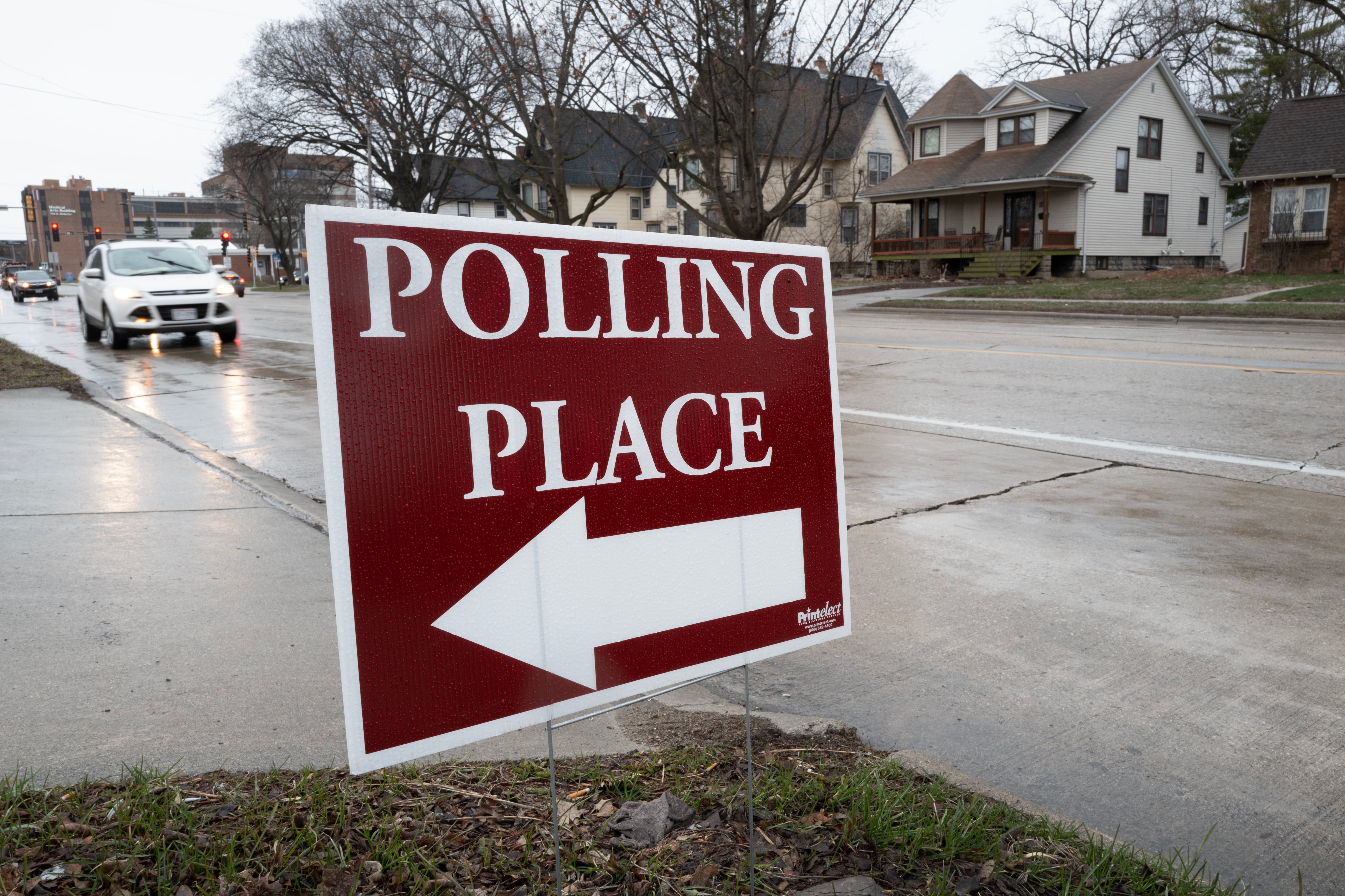 A red and white sign that reads "Polling Place" on the side of the road with houses and a car in the background.