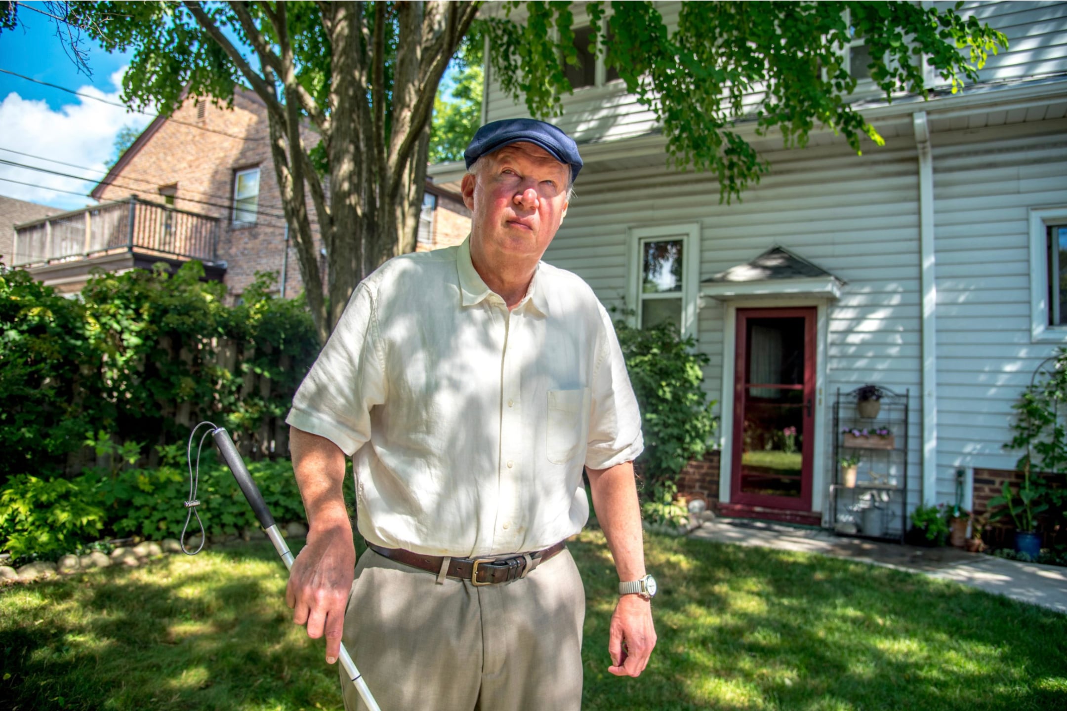 A man wearing a blue hat, a white shirt and holding a walking stick as a mobility device, stands in his green backyard with a large tree and a house in the background.