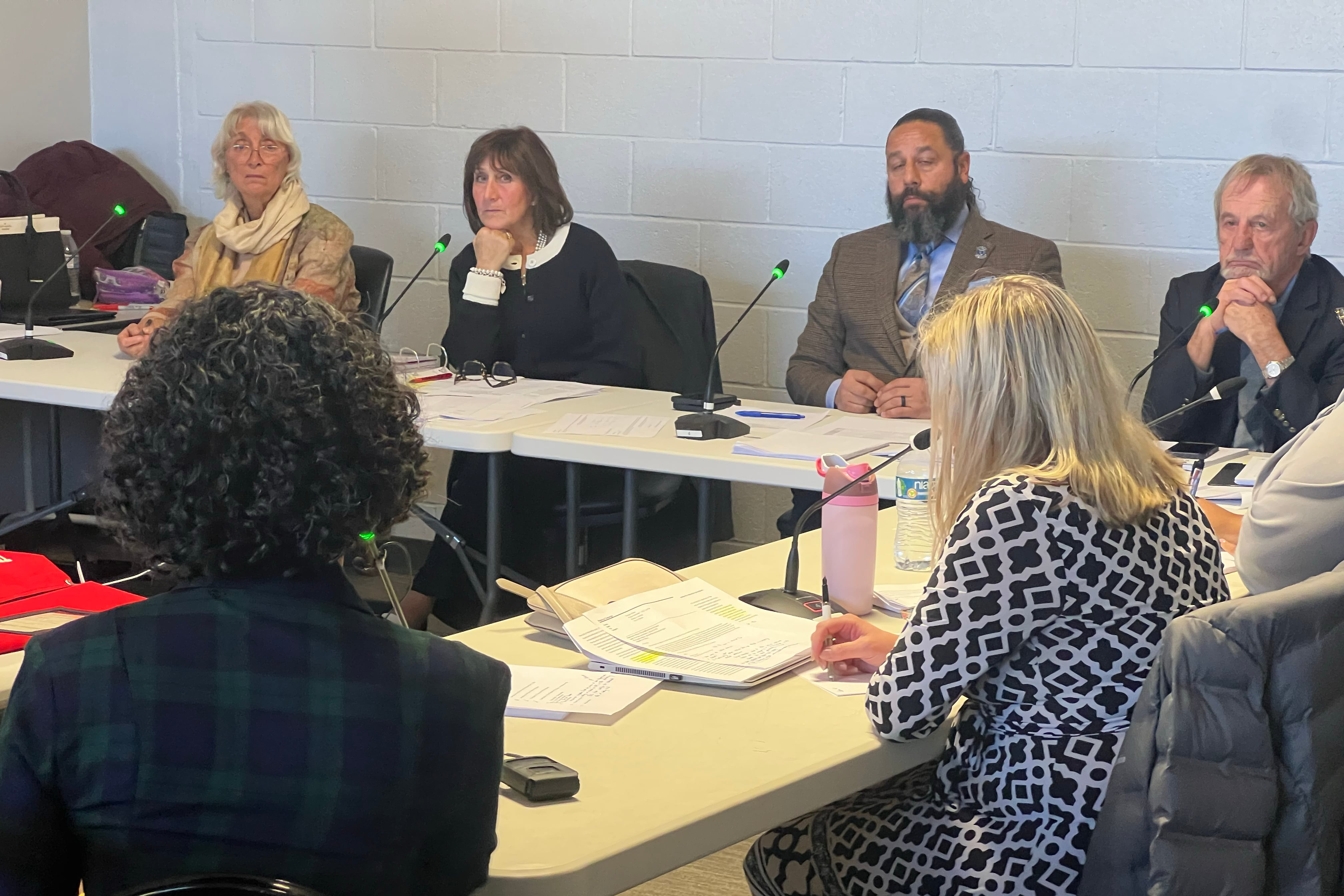 A photograph of a group of people wearing business clothing sit around folding tables with paper and microphones in front of each of them.