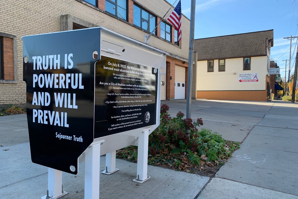 A ballot drop box located outside of a tan brick building with an American flag on the side.