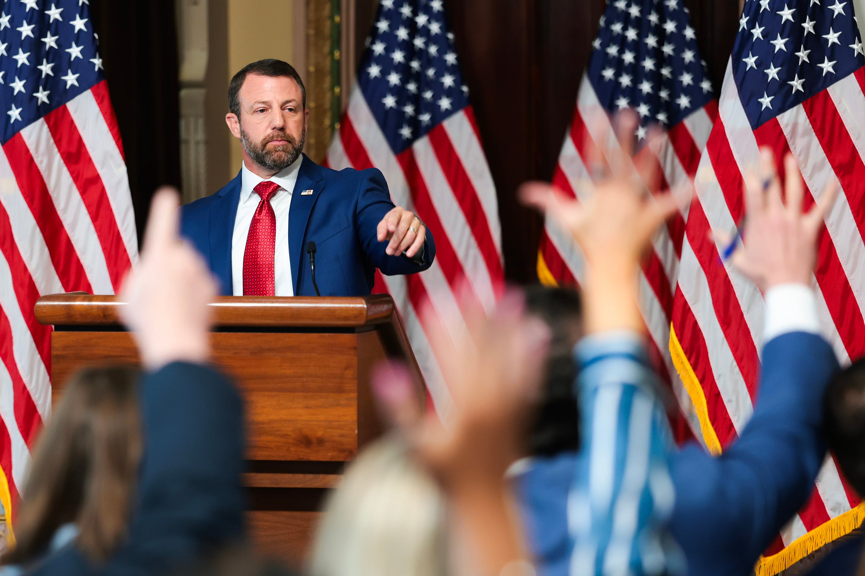 A white man in a suit and tie points to someone in a crowd to ask a question during a press conference. There are a row of blurry hands in the foreground and a row of American flags behind the man.