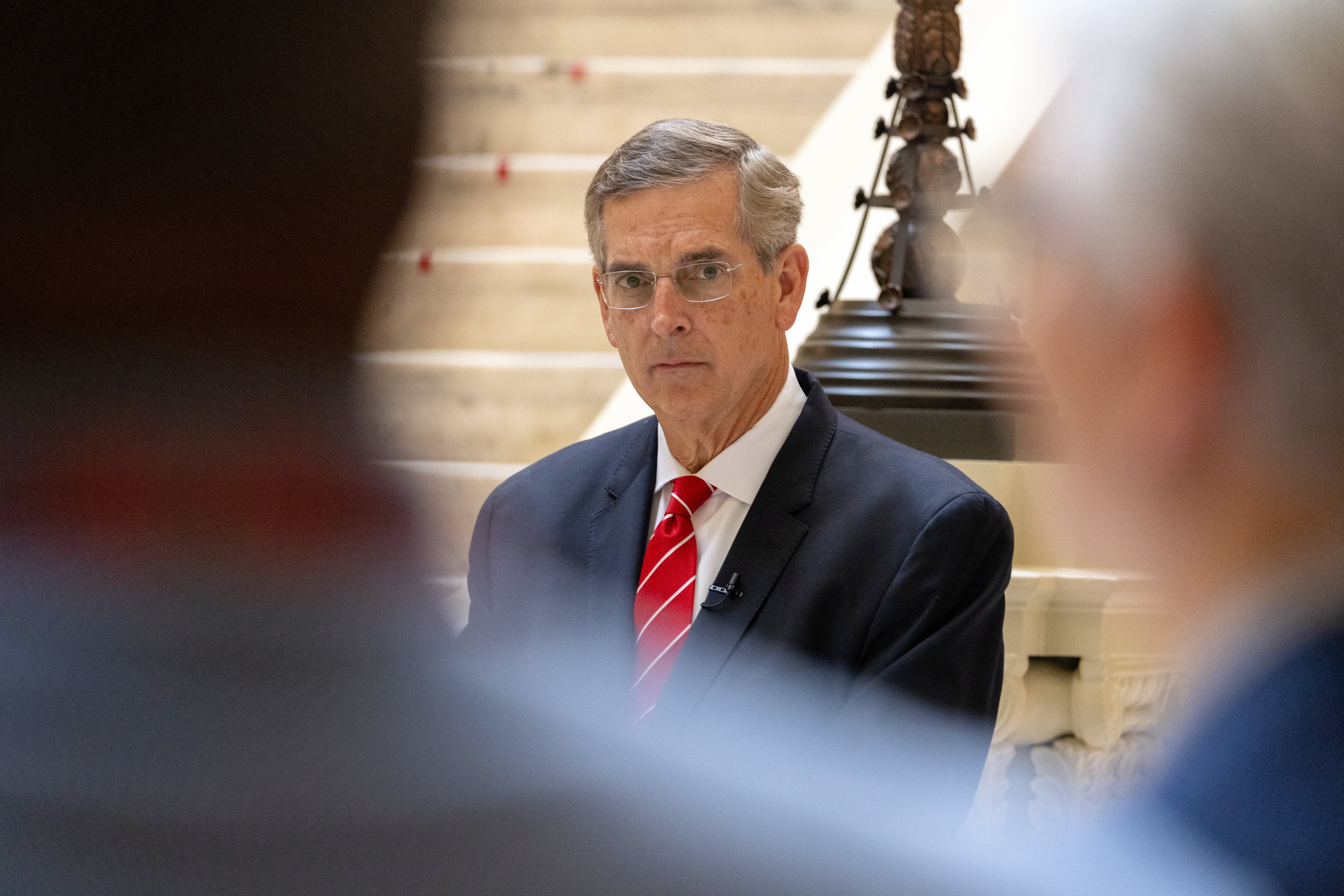 A man in a dark suit and red tie and wearing glasses stands in between two people who are in the foreground.