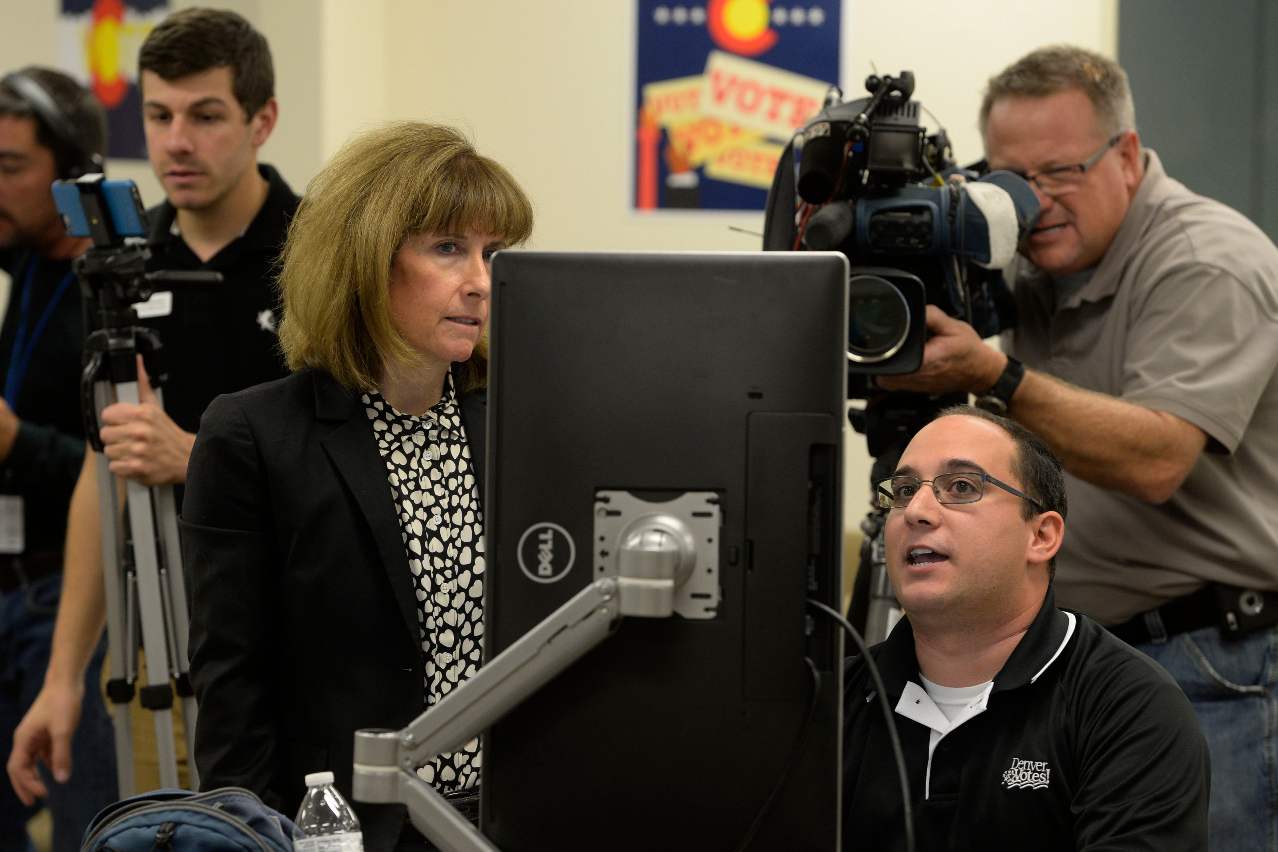 A woman stands next to a man seated before a computer screen as they both look at it, with a man holding a television camera in the back, capturing the scene.