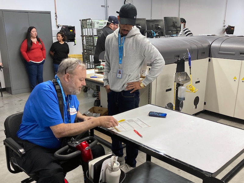 A man in a blue shirt and sitting in an electric powered chair works at a table while another man wearing a white sweater and a black hat stands next to him with other people in a warehouse looking room in the background.