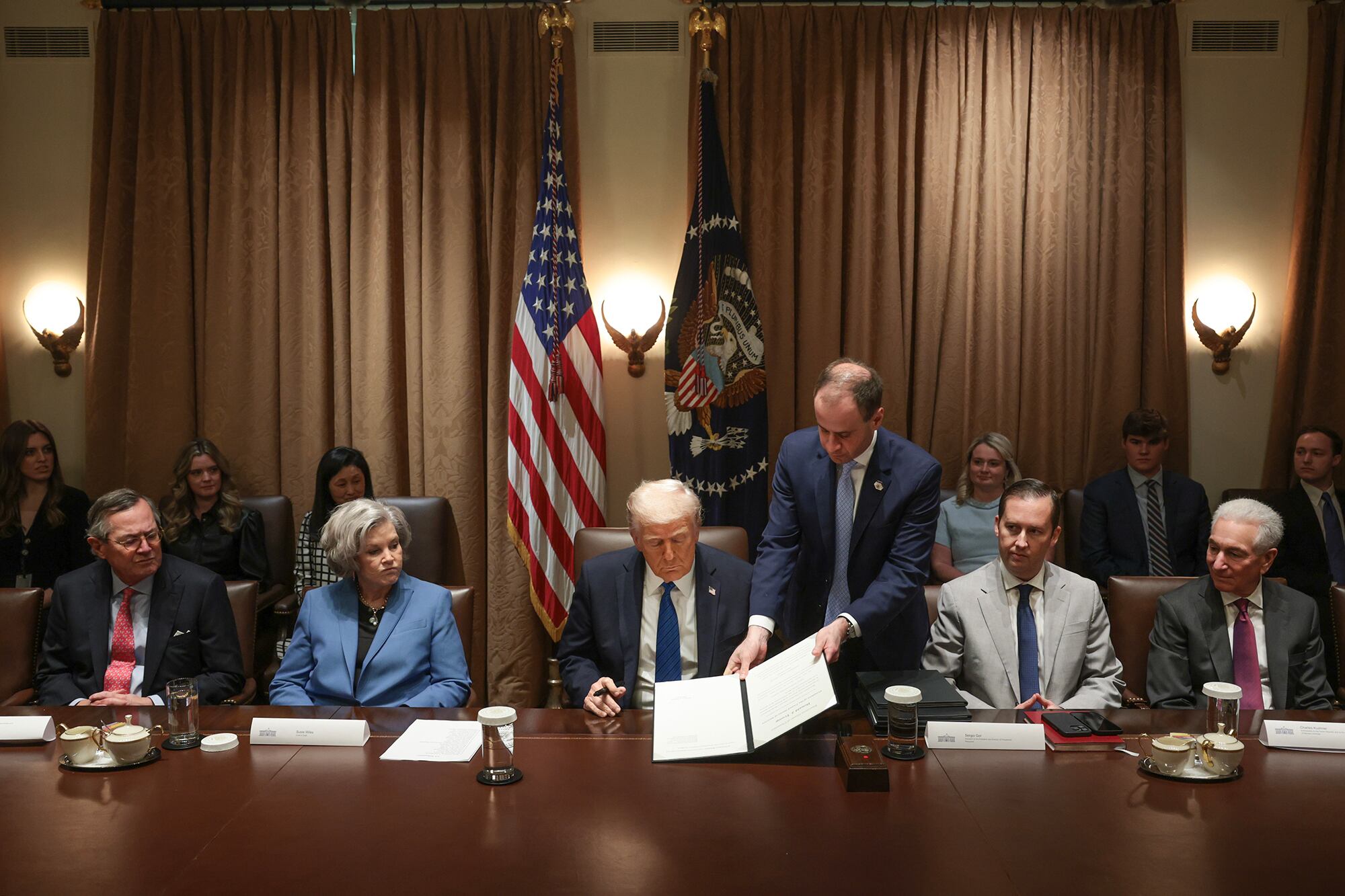 Five people in suits sit at a long wooden table in the White House while one man stands next to Donald Trump in the middle and a row of people and two flags are in the background.