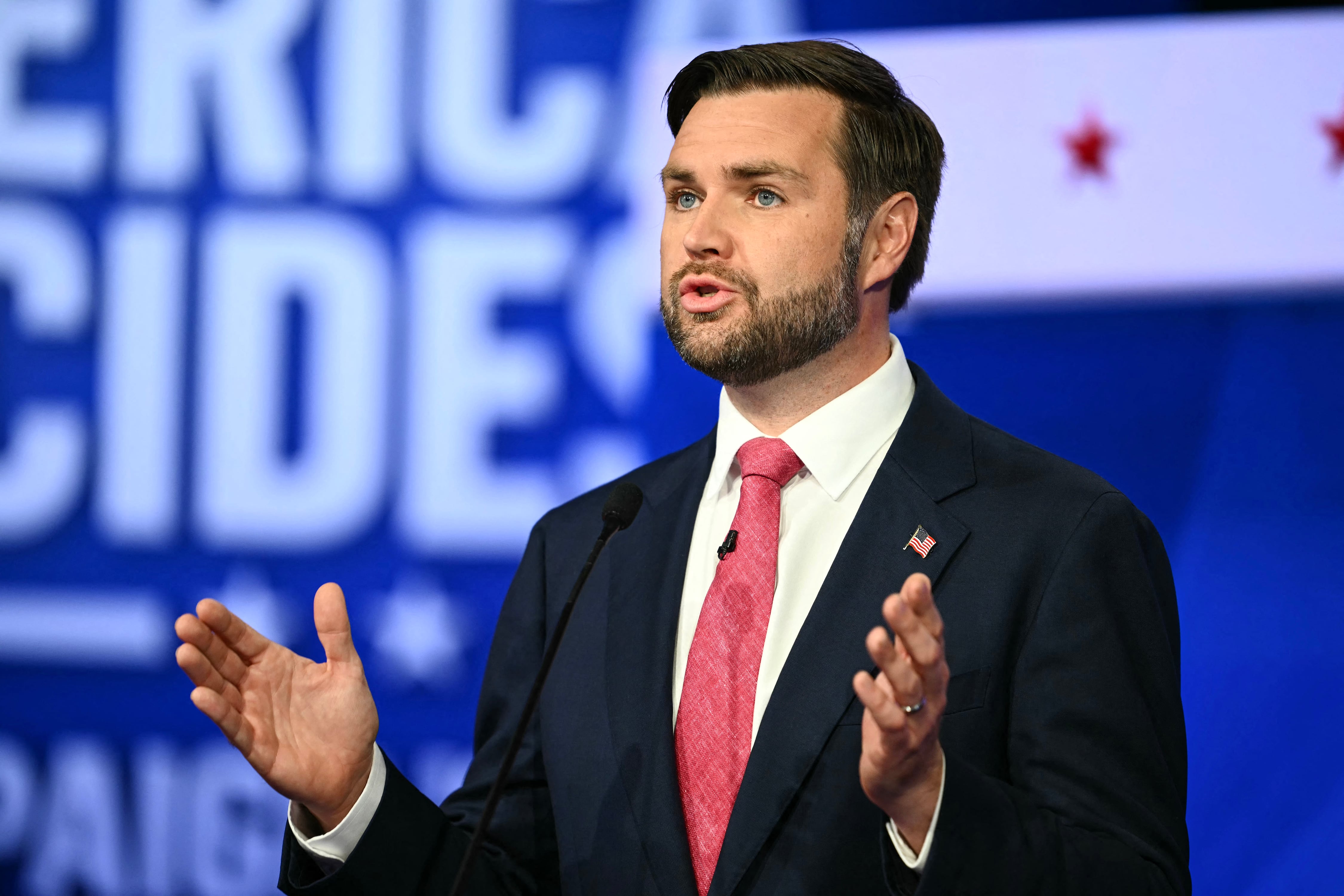 A man in a dark suit and a wearing a red tie holds his hands in the air while talking and a white and blue background.