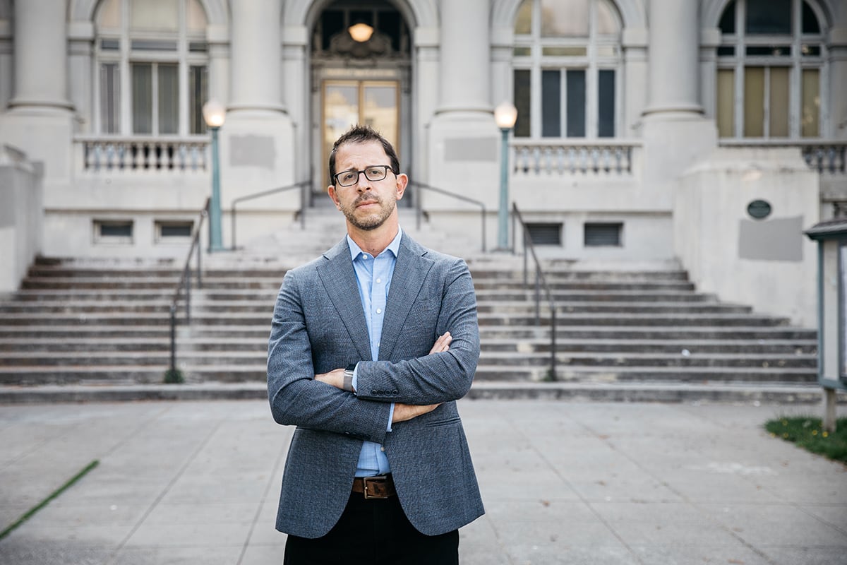 A man wearing black glasses, a light blue shirt, and grey sportscoat stands in front of a flight of stone steps, with his arms crossed in front of him. 