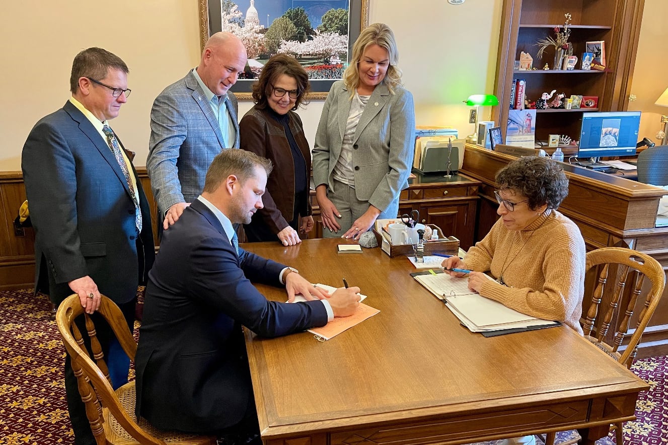 Two people sit at a wooden table filling out paperwork while four adults stand to the side watching.