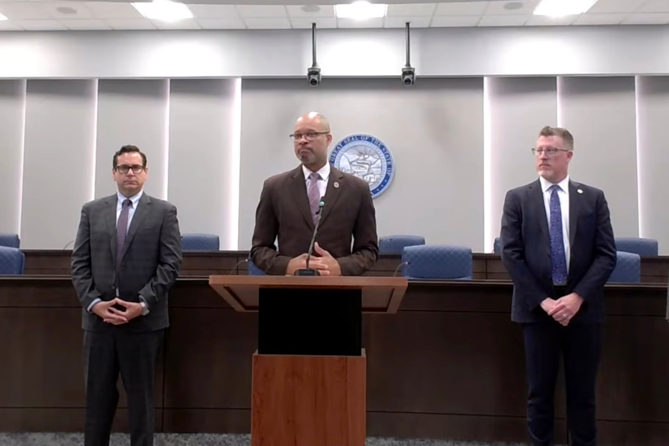 Screengrab of a press conference showing three men in suits standing side by side.