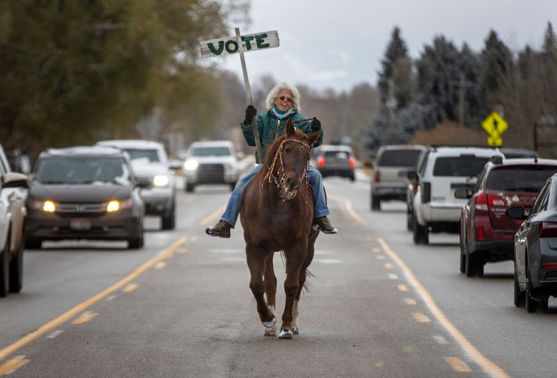 A woman rides a horse through a busy street holding a vote sign.