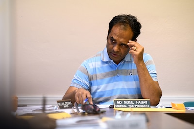 A man in a blue and white striped shirt with dark hair holds his head in his hand while sitting at a table in a conference room.
