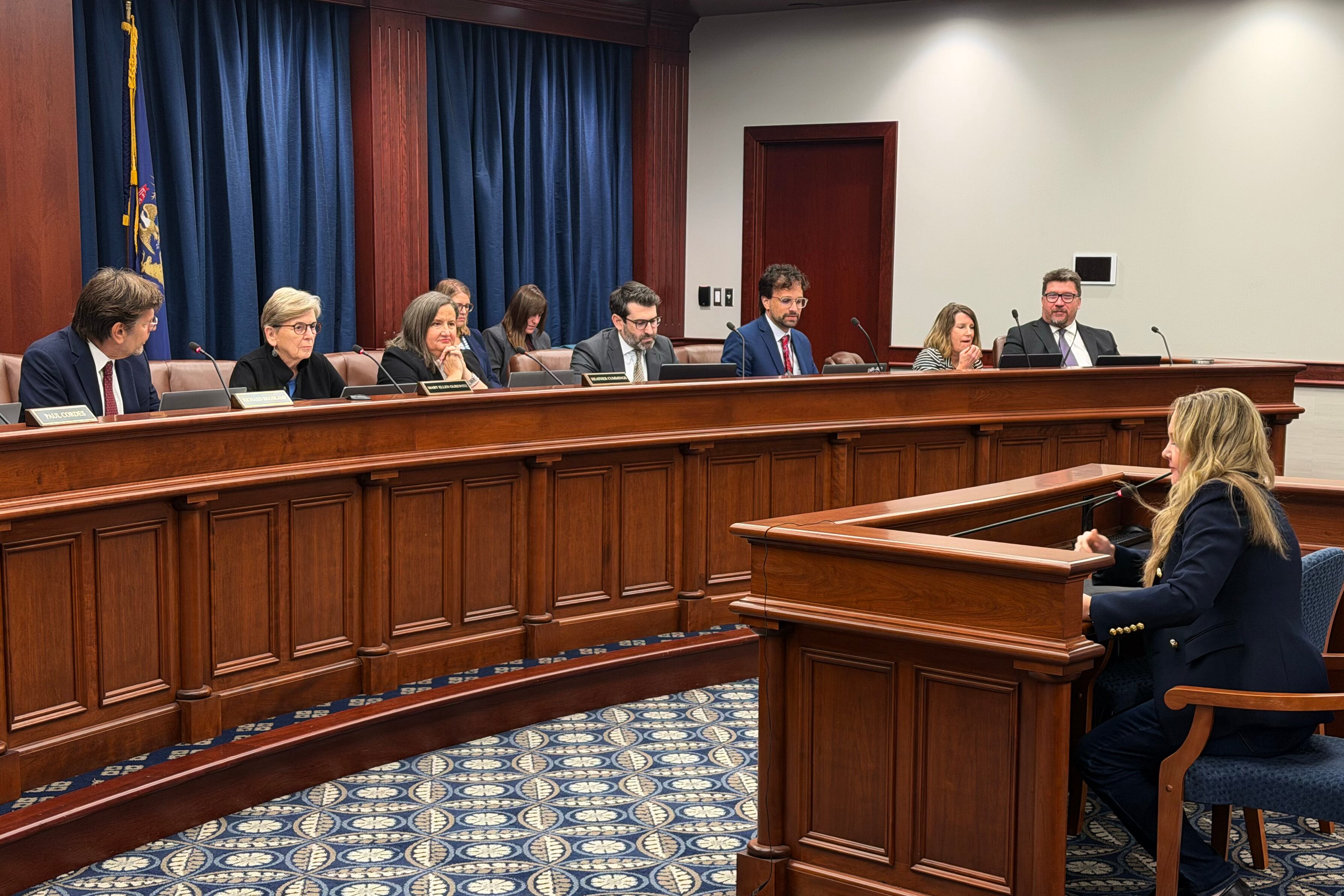 A group of adults sit at a long wooden table in a large state conference room.