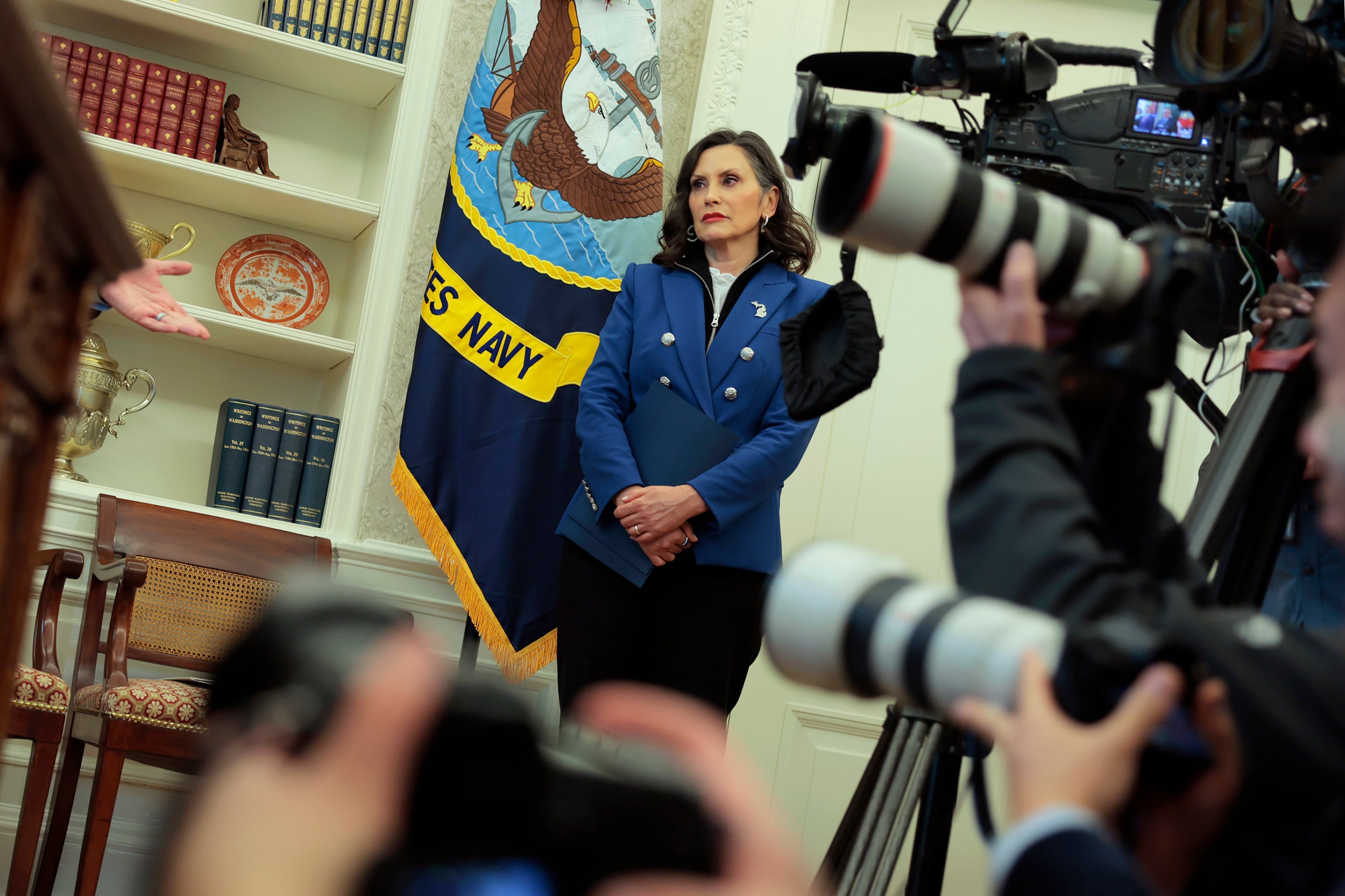 A photograph of a white woman in a suit stands in the background while photojournalists' cameras make a circle in the foreground.