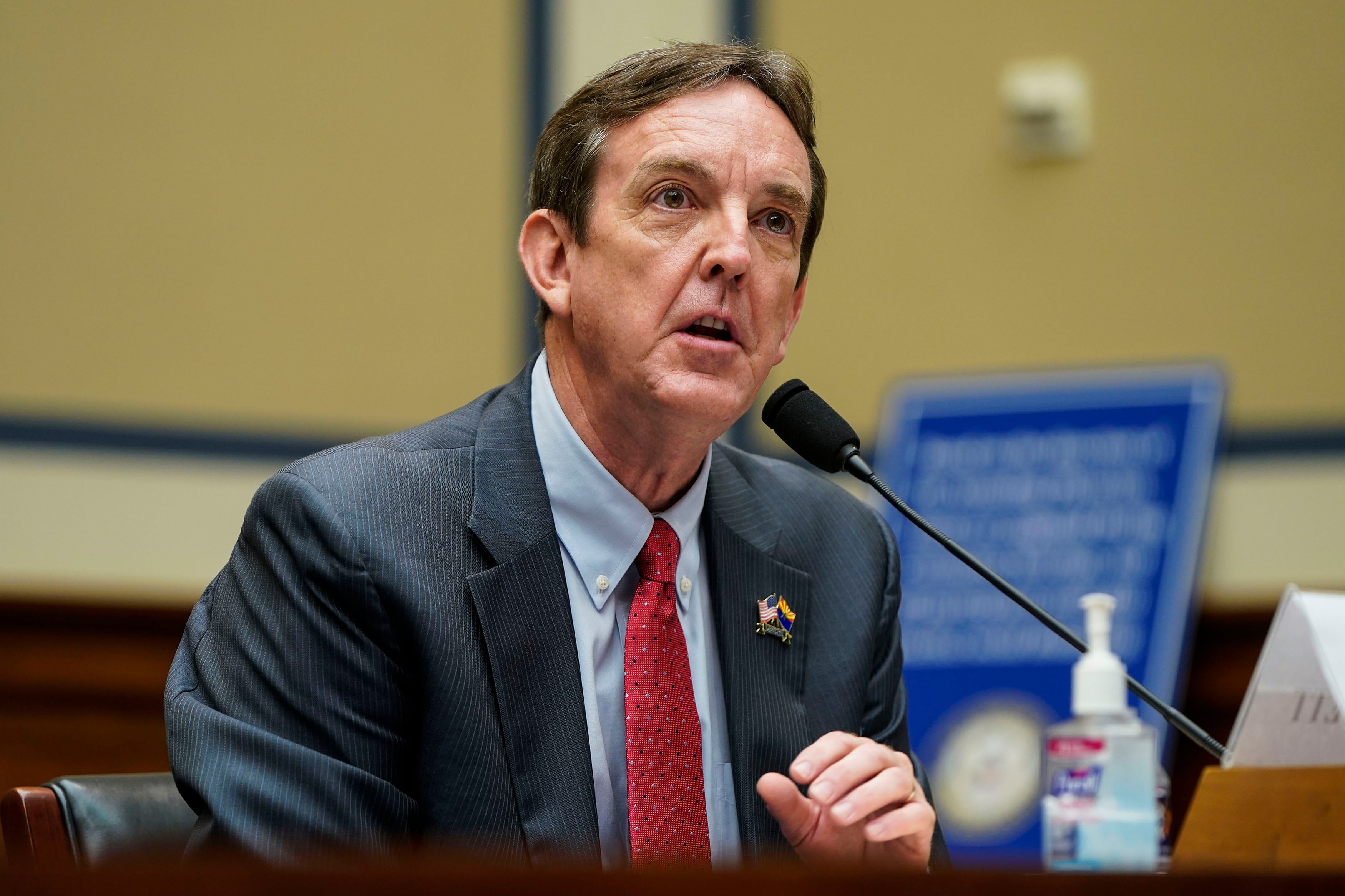 A man with short dark hair and wearing a dark suit and red tie sits at a wooden table and speaks into a microphone.