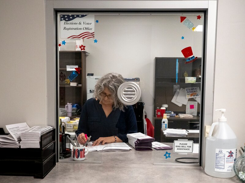 A woman with short gray hair works behind a glass window.