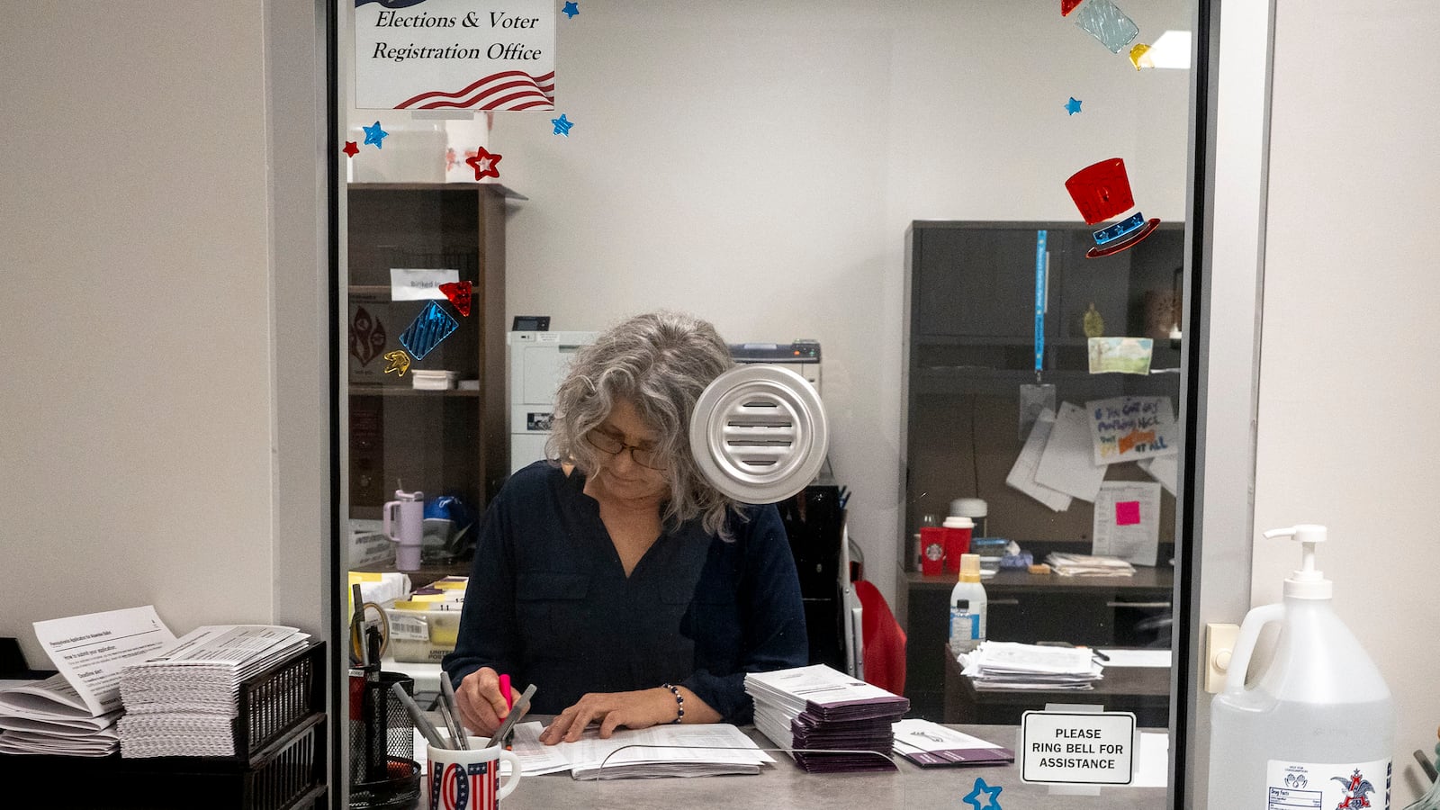 A woman with short gray hair works behind a glass window.