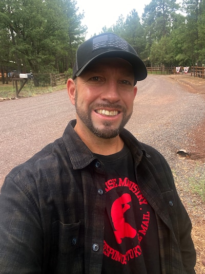 A white man in a black hat and a plaid shirt stands outside posing for a portrait.