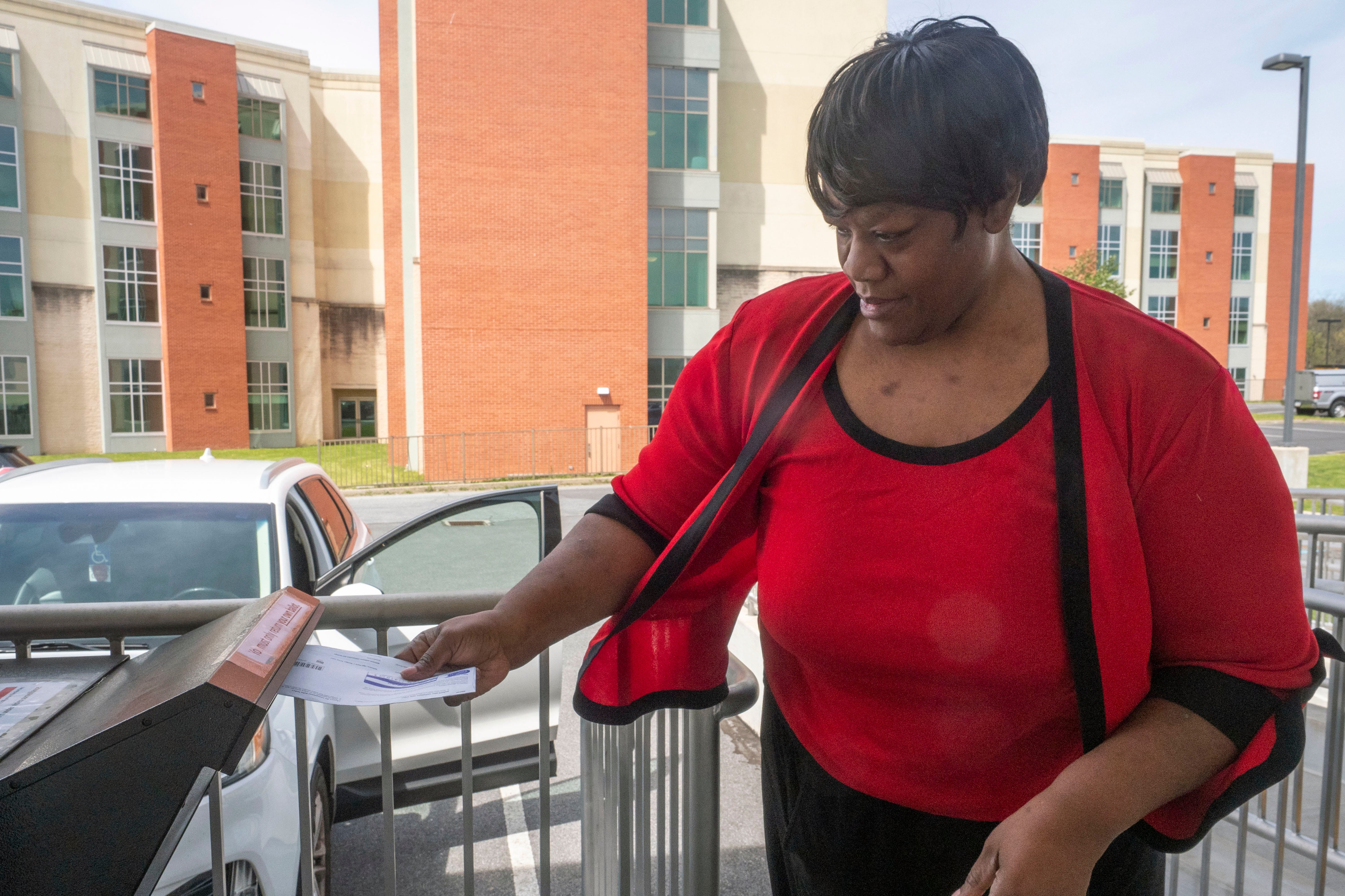 A person wearing a read blouse places a ballot into a box with a car and orange brick building in the background.