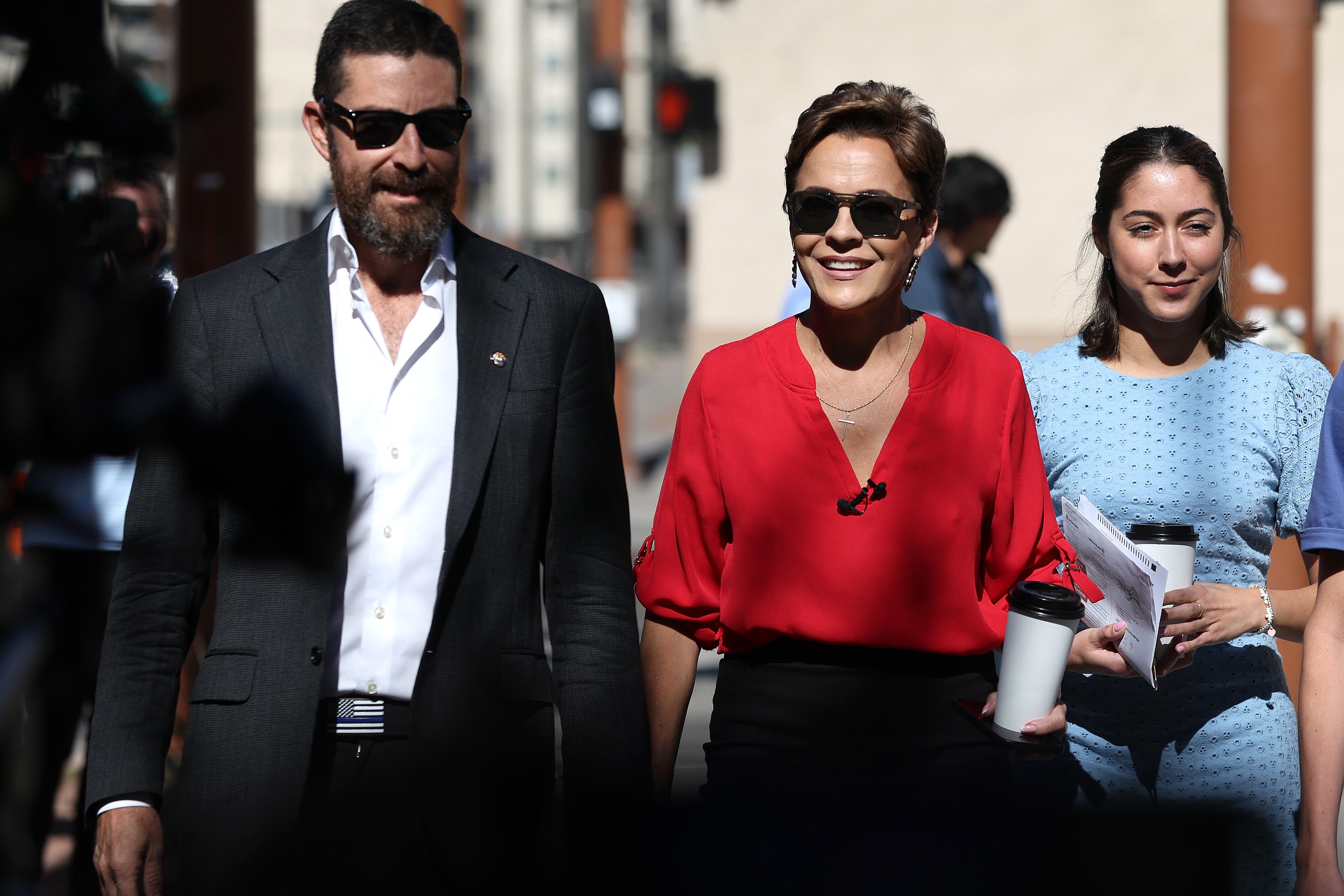 A bearded man in a dark suit and sunglasses holds the hand of a woman wearing a red blouse and sunglasses and holding a coffee cup as they walk.