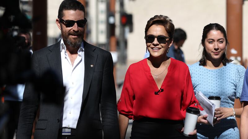 A bearded man in a dark suit and sunglasses holds the hand of a woman wearing a red blouse and sunglasses and holding a coffee cup as they walk.
