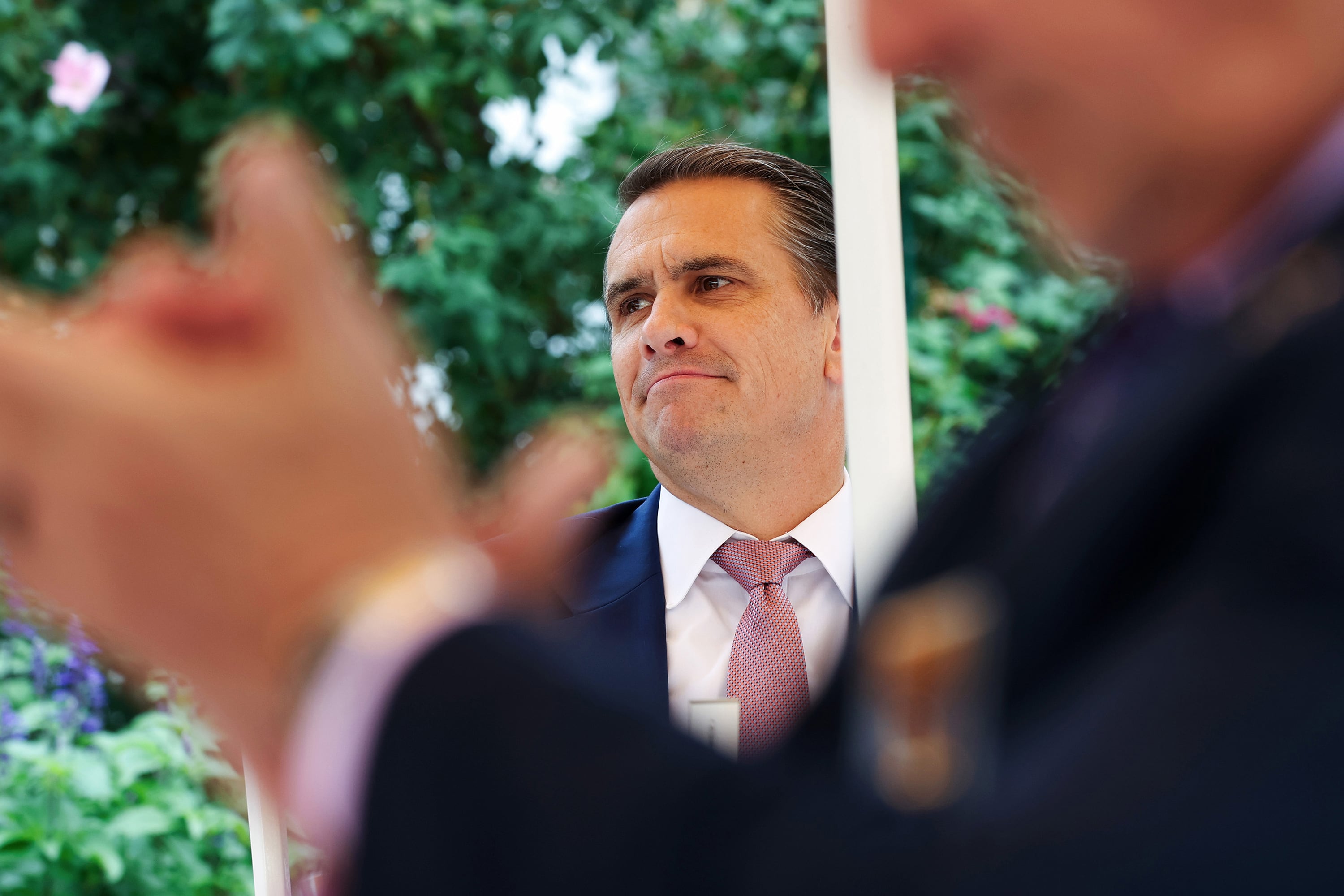 A white man in a suit and tie stands outside against some greenery while a man claps in the foreground.