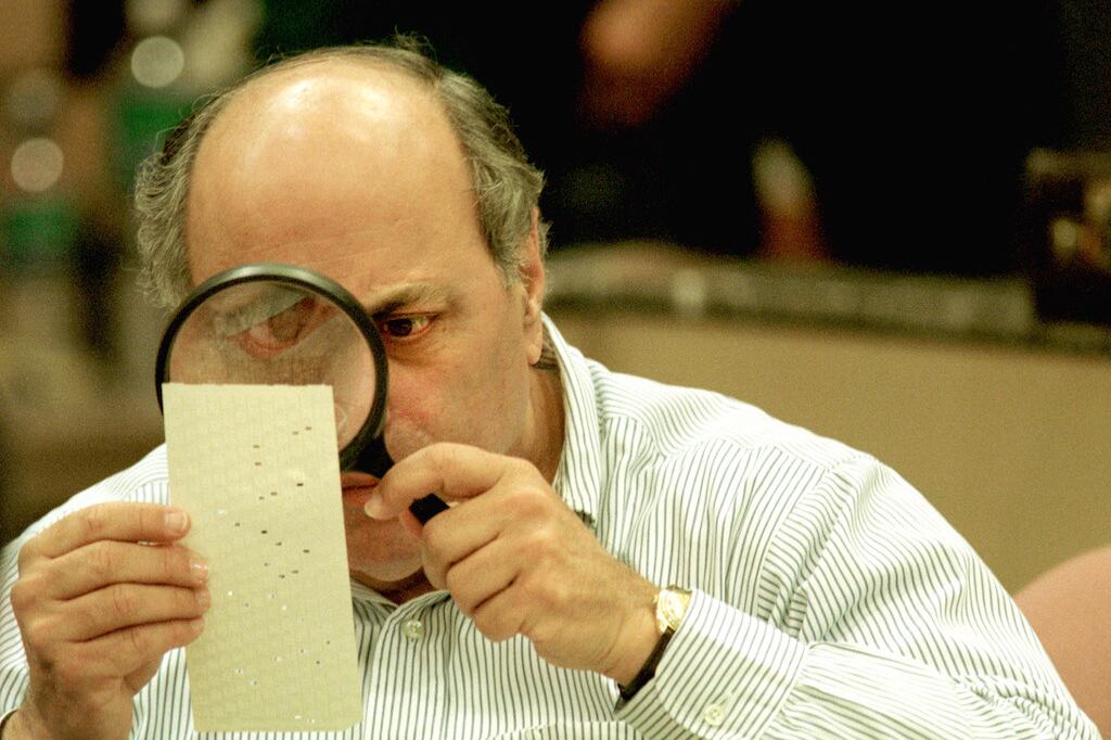 A man holds a magnifying glass to a punch card ballot.