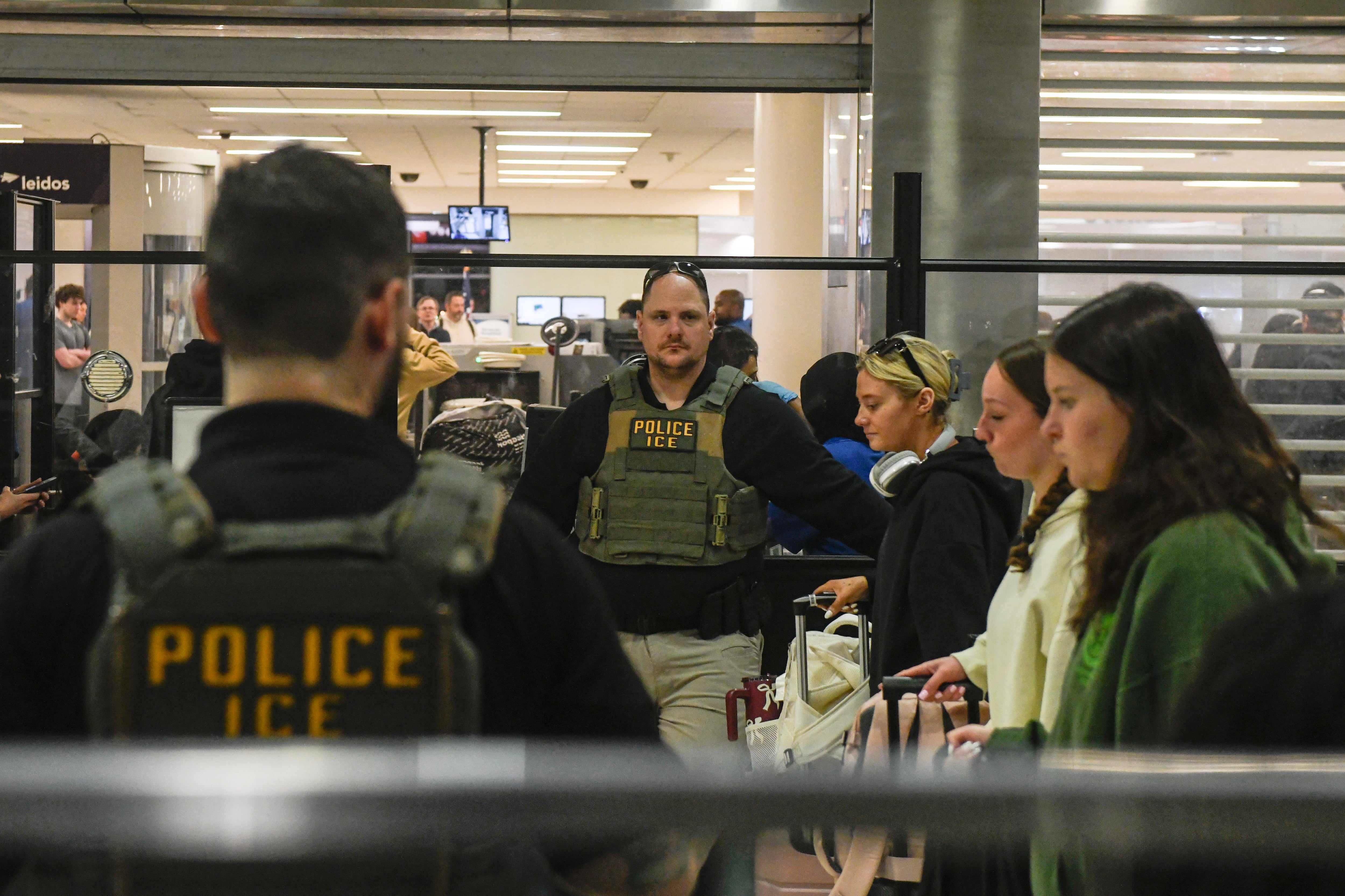 Two ICE agents watch over people at an airport.