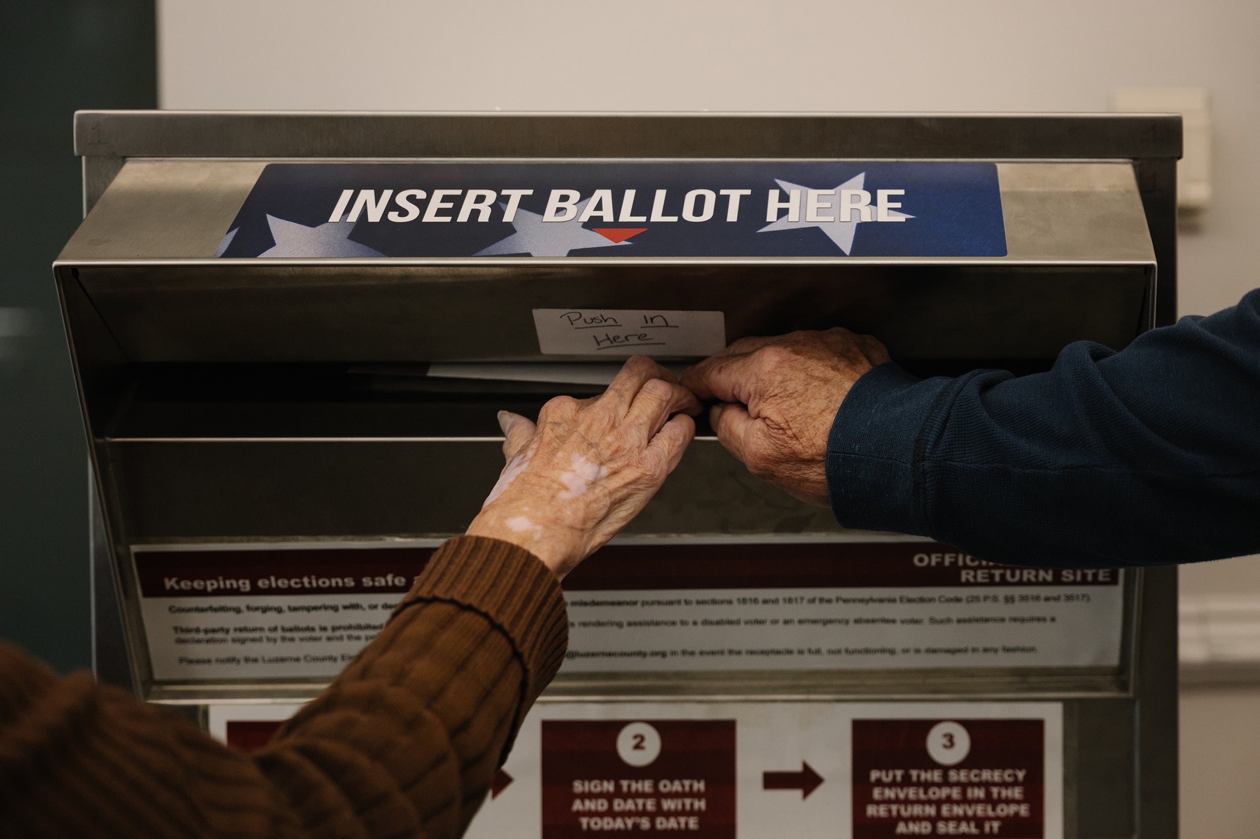 A photograph of two hands pushing a mail in ballot into a return ballot box.