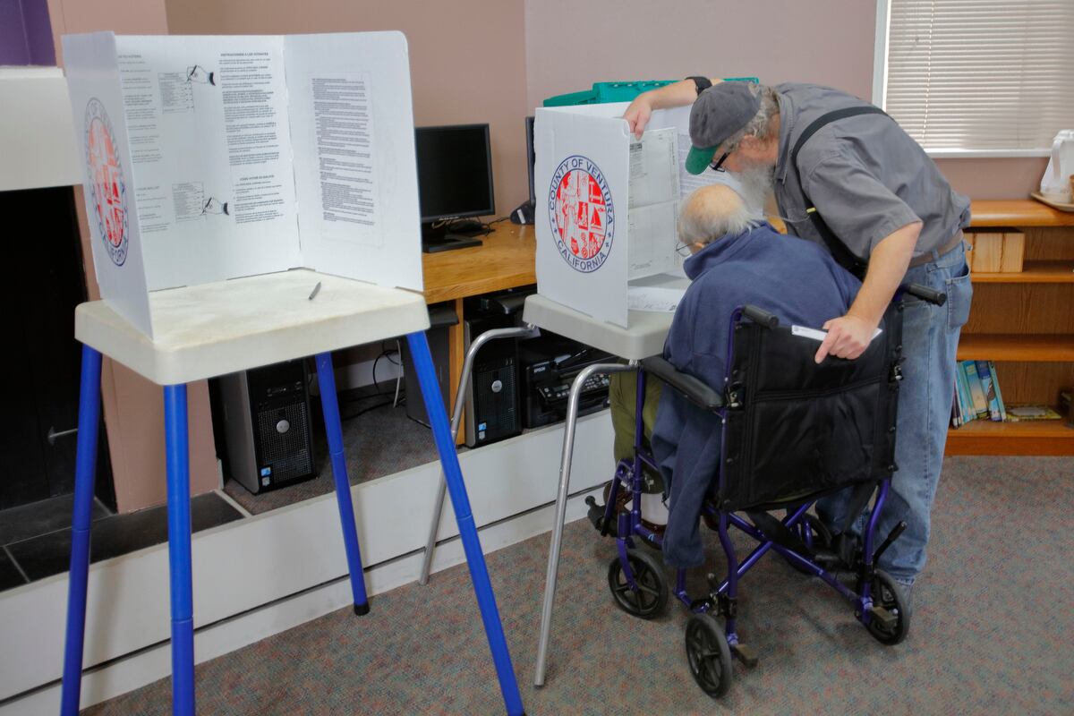A man in a baseball cap leans over next to a man in a blue coat sitting in a wheelchair, looking at a blown-up ballot.&nbsp;