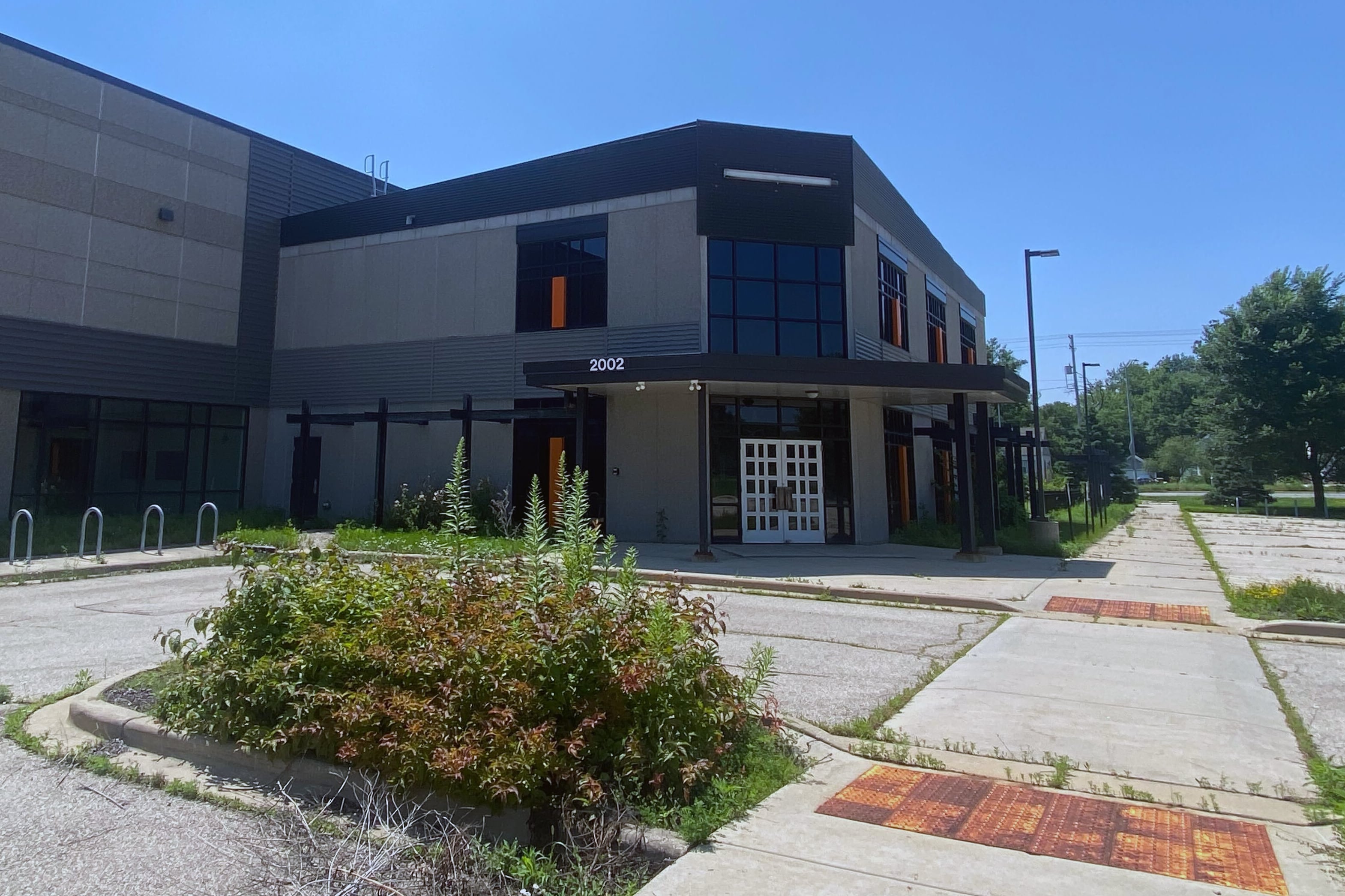 A large building with a shrub in the foreground a sidewalk on the right side and a blue sky in the background.
