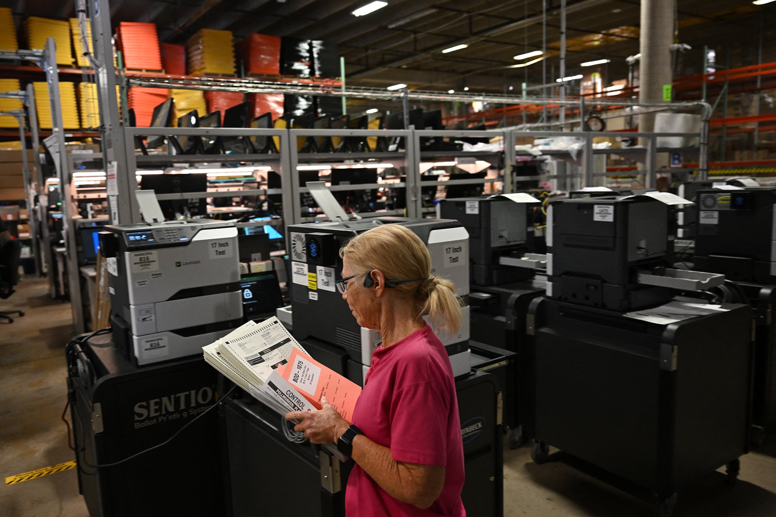 A photograph of a white woman in a red t-shirt holding a stack of papers walking by a large room full of voting machines.