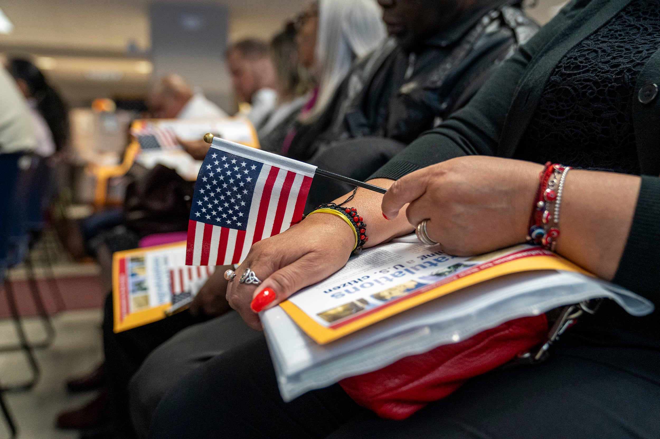 A photograph of a person's hand holding a small American flag and papers in a large room with other people in the background.