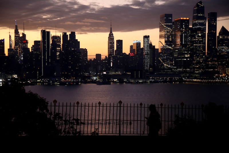 The sun rises behind the skyline of Manhattan with a river in the middle and a person walking along a fence line in the foreground.