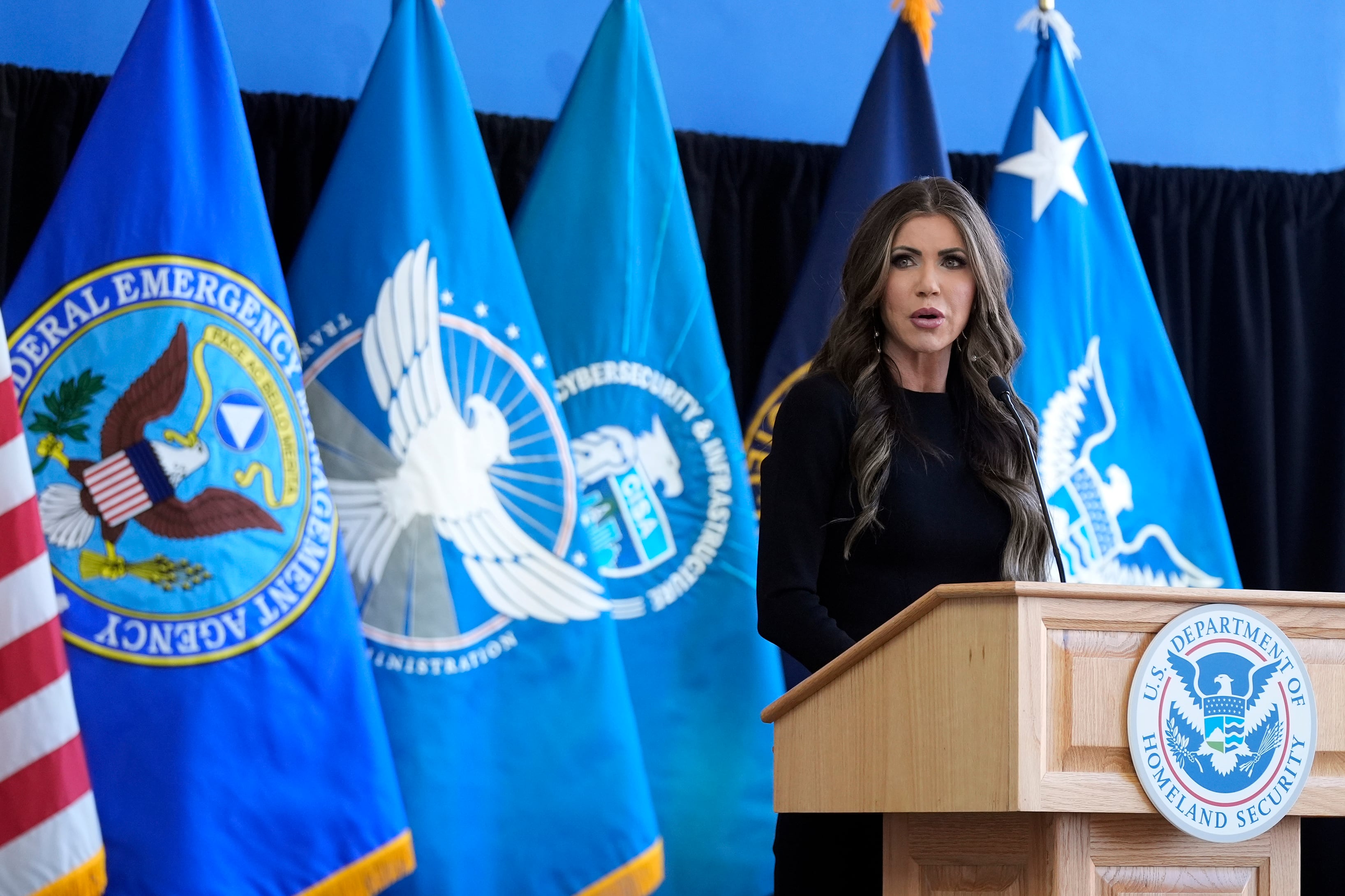 A woman with long dark hair stands behind a podium and in front of a row of flags.