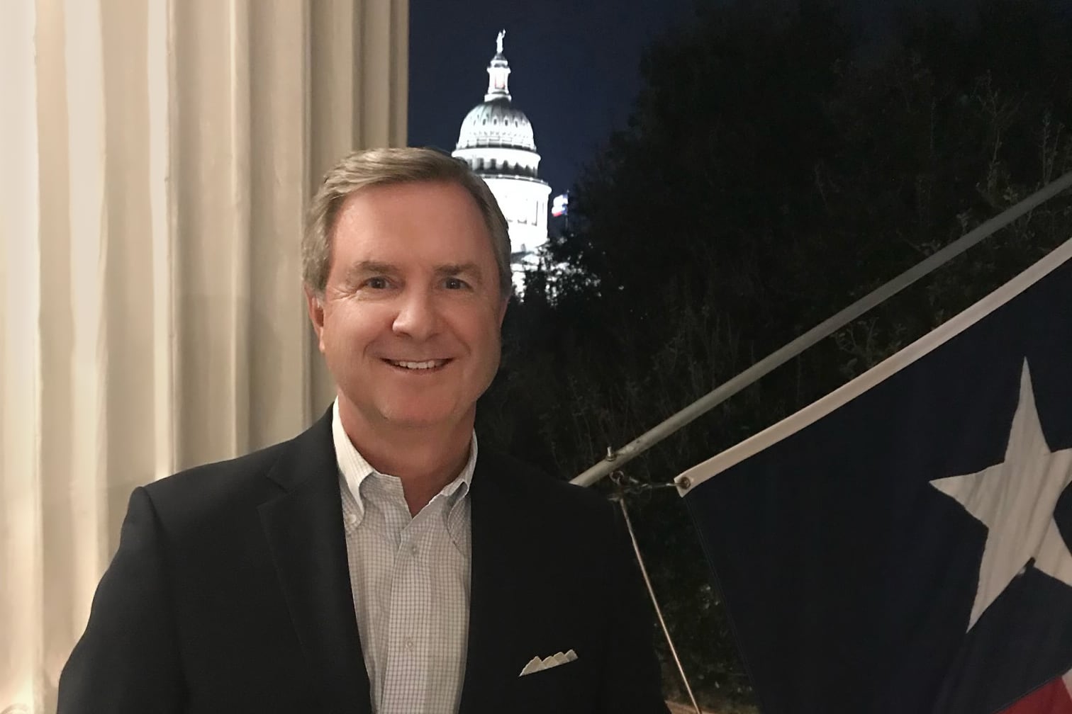 A white man in a dark suit jacket poses for a portrait on a balcony with a Texas flag on the right of the frame and the State Capitol building in the background.