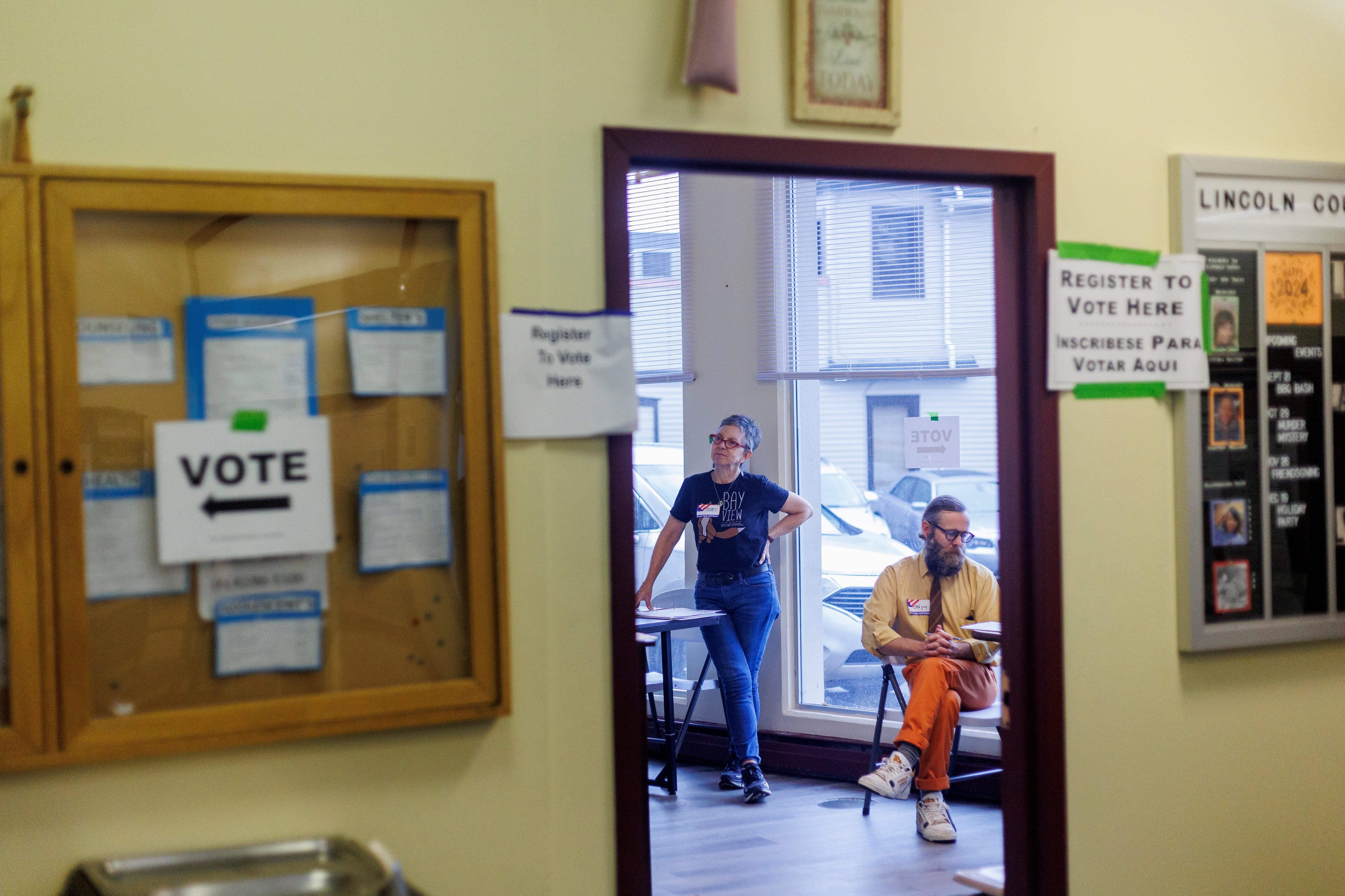 Two people stand in the background of a polling location with a wall and doorway in the foreground.