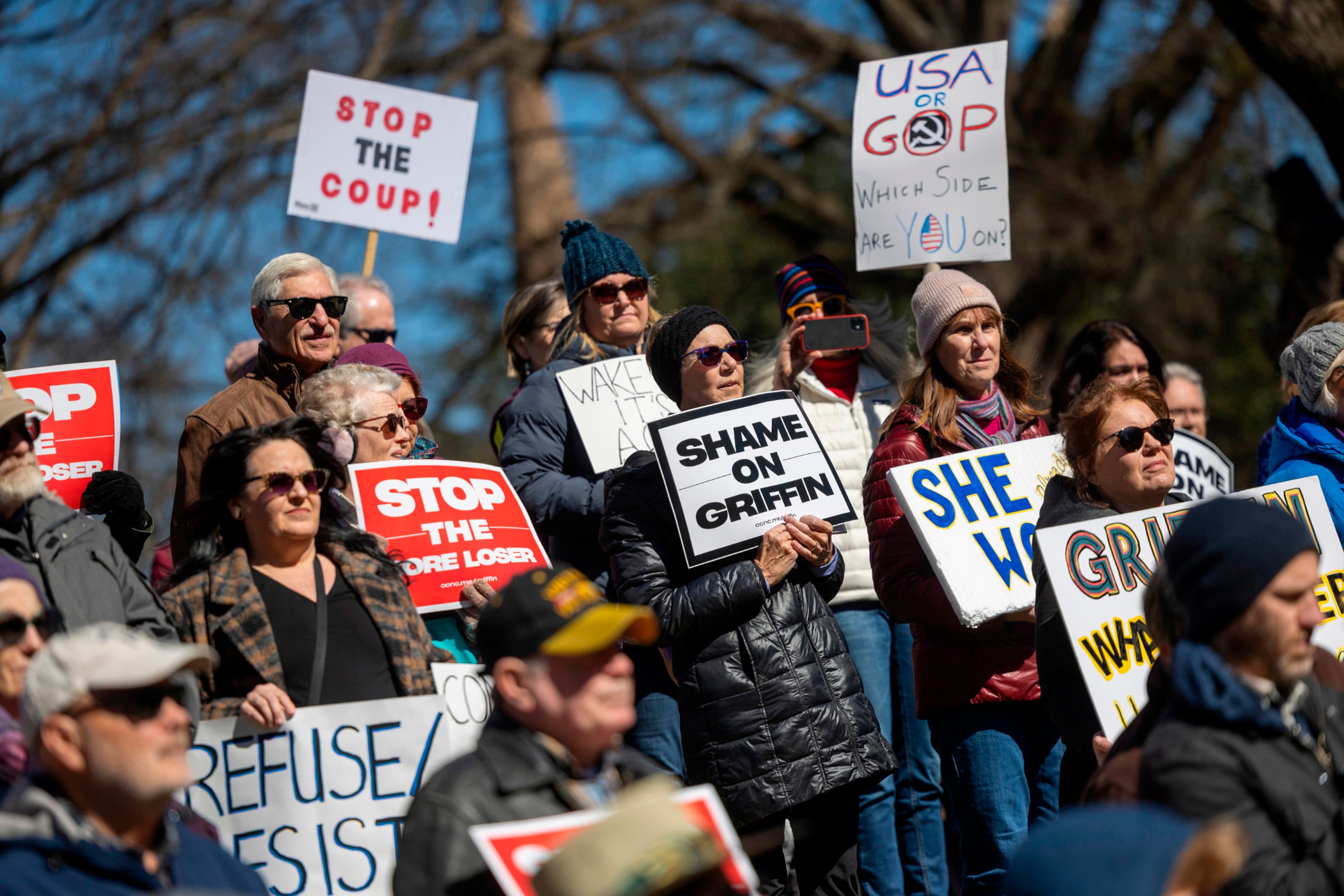 A crowd of people stand outside holding signs while protesting on a sunny day.