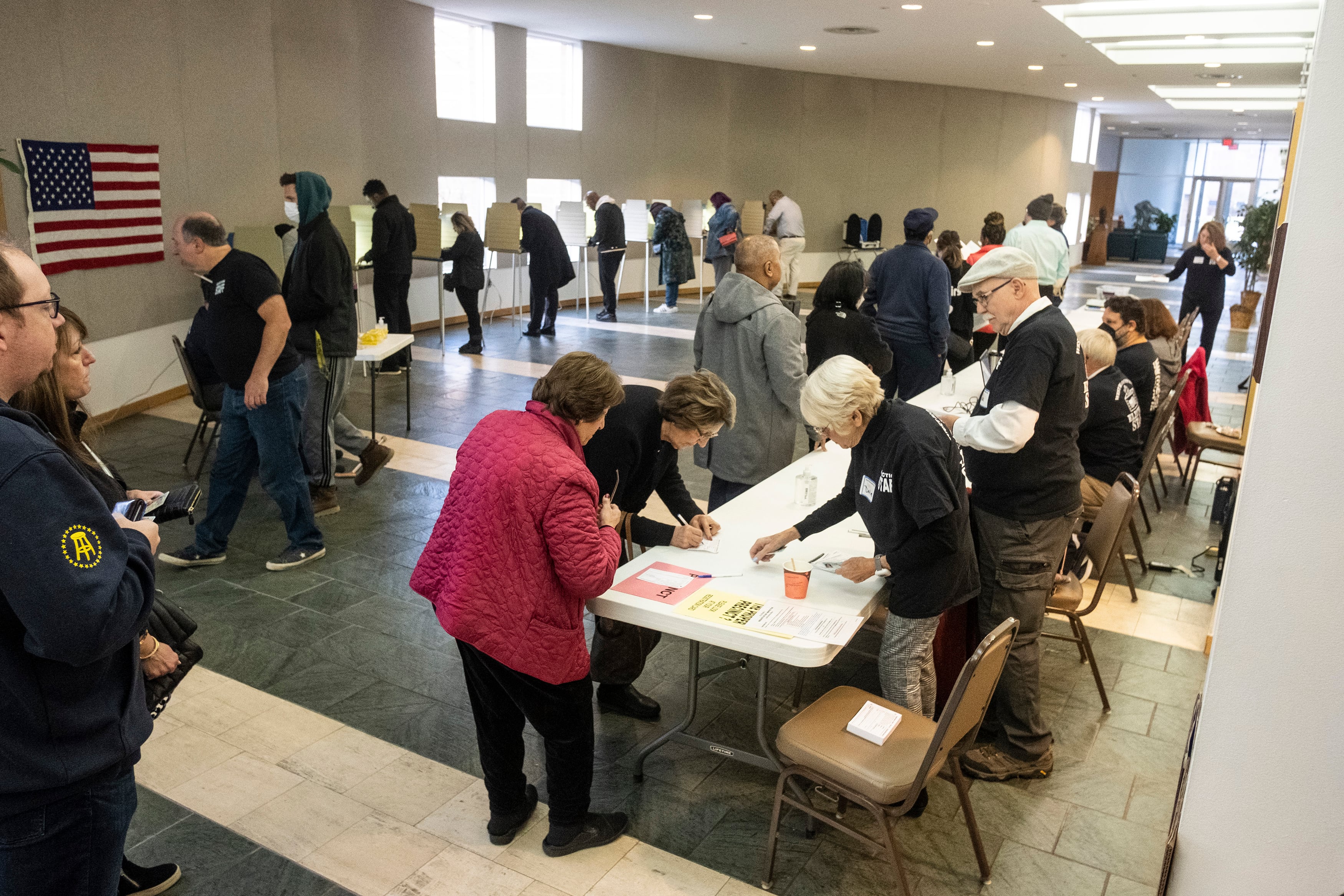 People line up at a table in a large room while workers speak to them and assist