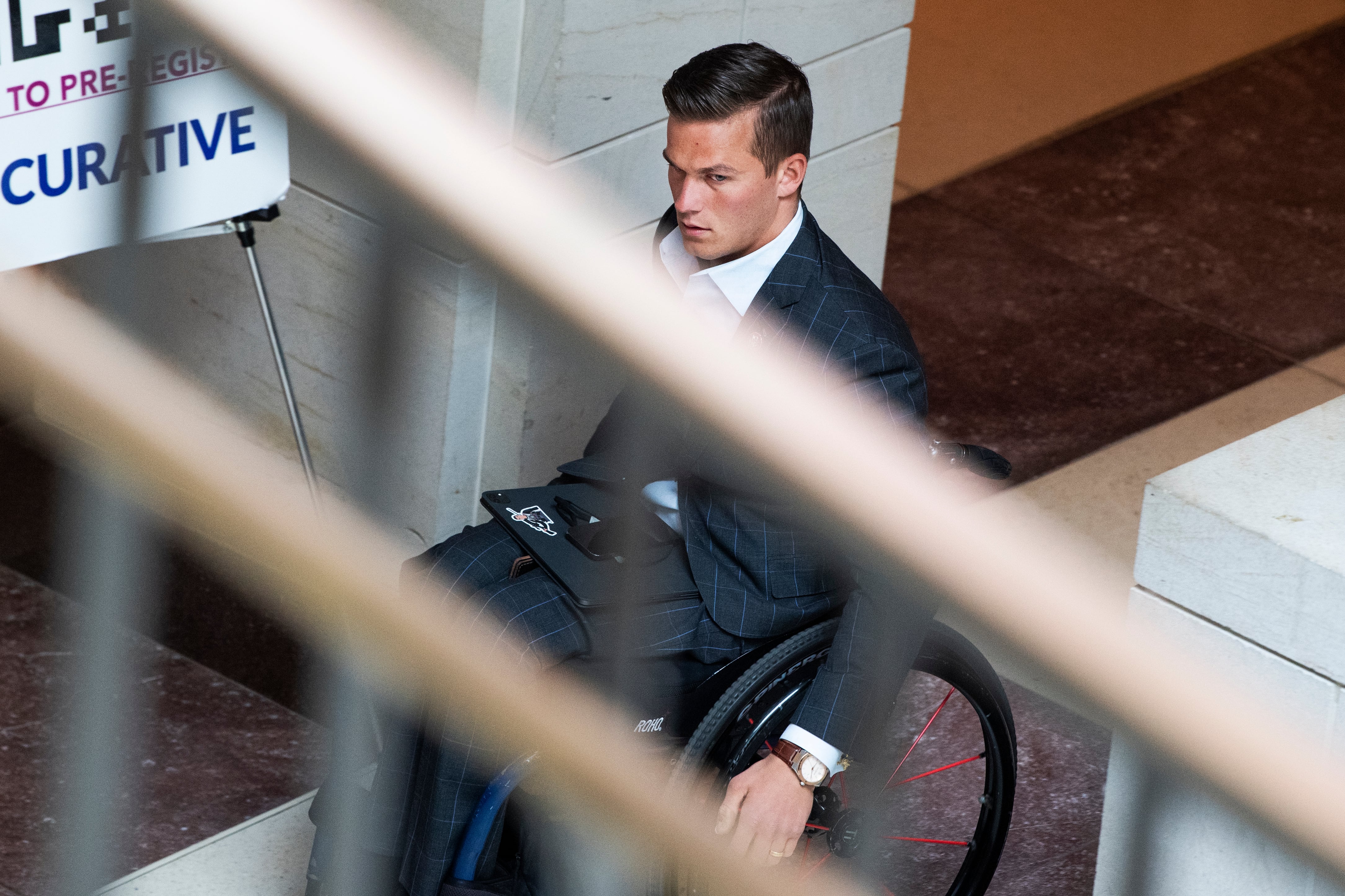 Madison Cawthorn, wearing a suit and moving his wheelchair down a hallway, viewed from above through the railing bars of a nearby staircase