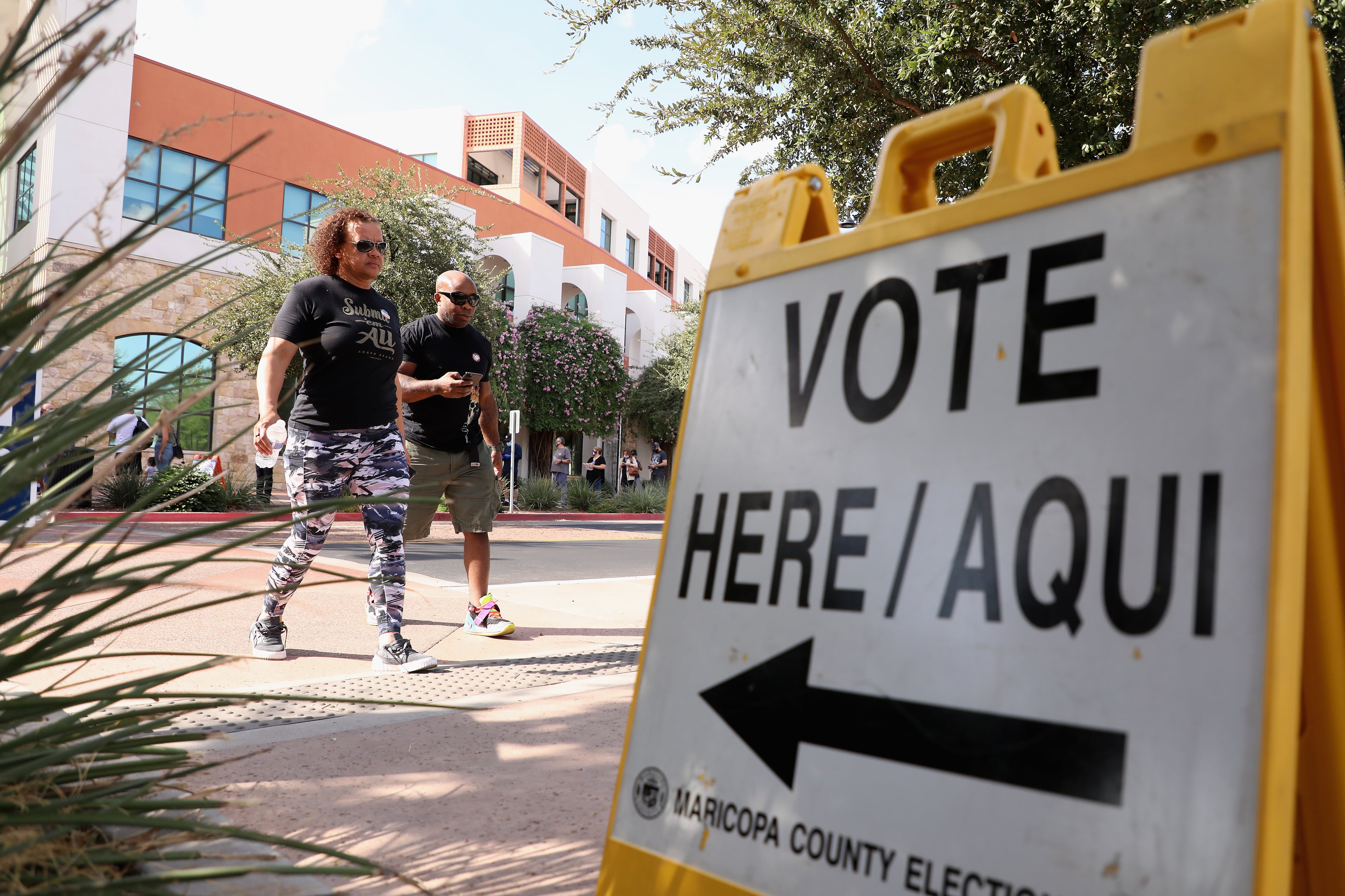 A woman and man cross a street in front of a yellow sign reading “Vote here / aqui”