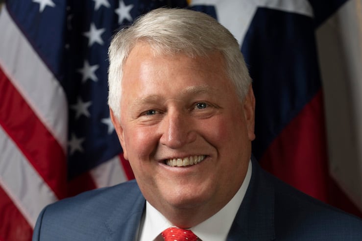 John Scott smiling and posing before a a U.S. flag and Texas flag