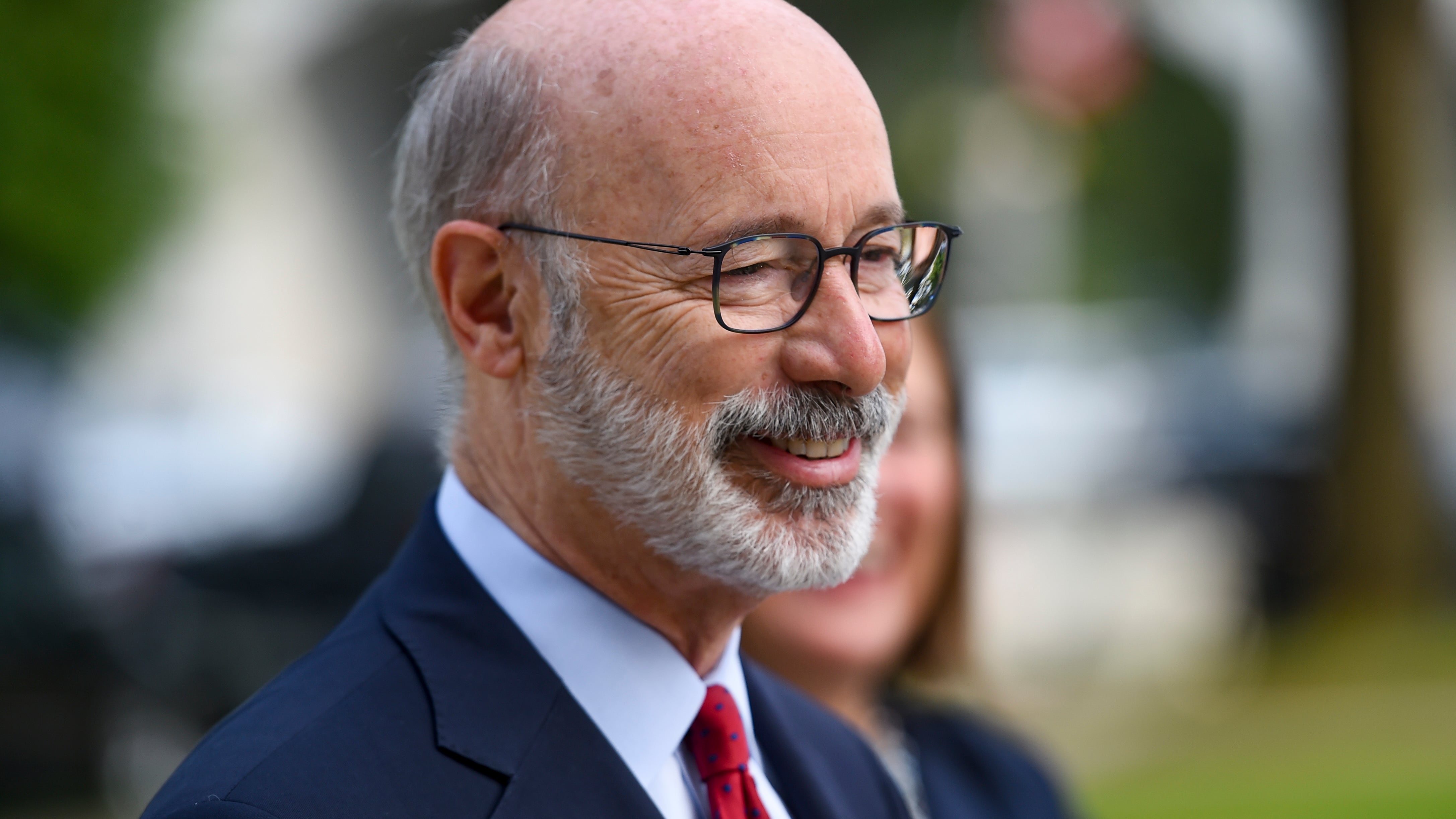 Closeup of Tom Wolf in profile, smiling and listening at an outdoor event, wearing a blue suit and red tie
