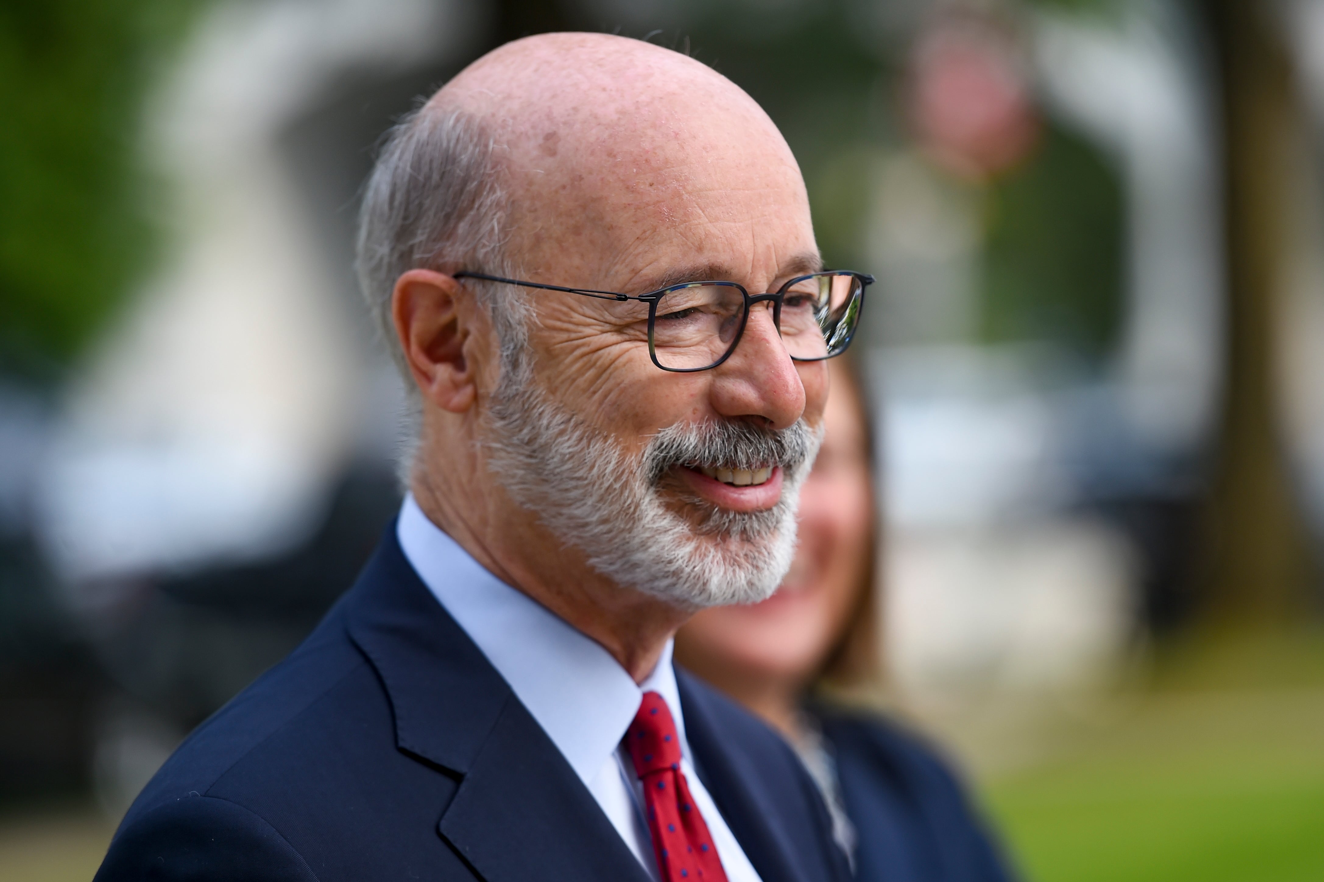 Closeup of Tom Wolf in profile, smiling and listening at an outdoor event, wearing a blue suit and red tie