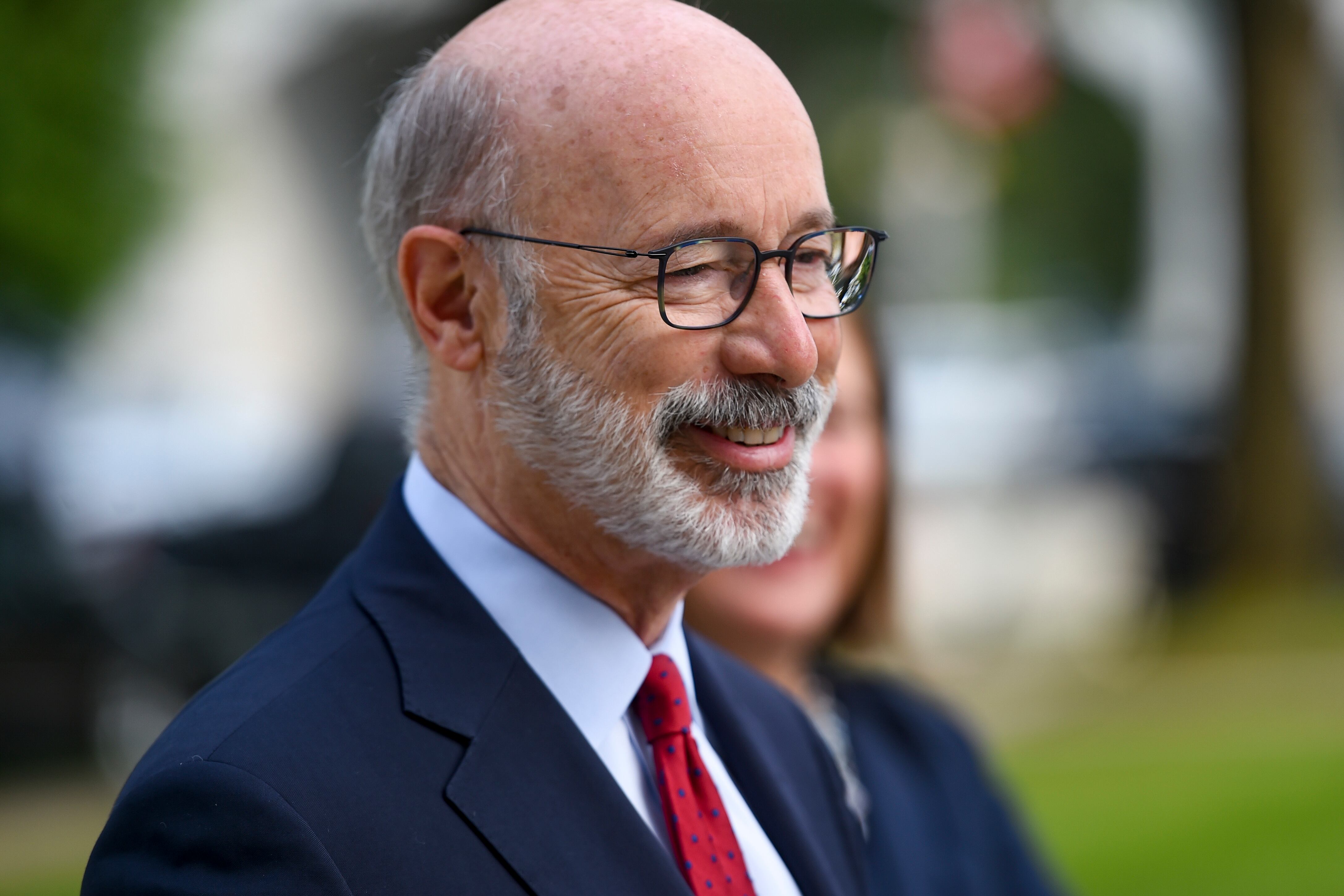 Closeup of Tom Wolf in profile, smiling and listening at an outdoor event, wearing a blue suit and red tie