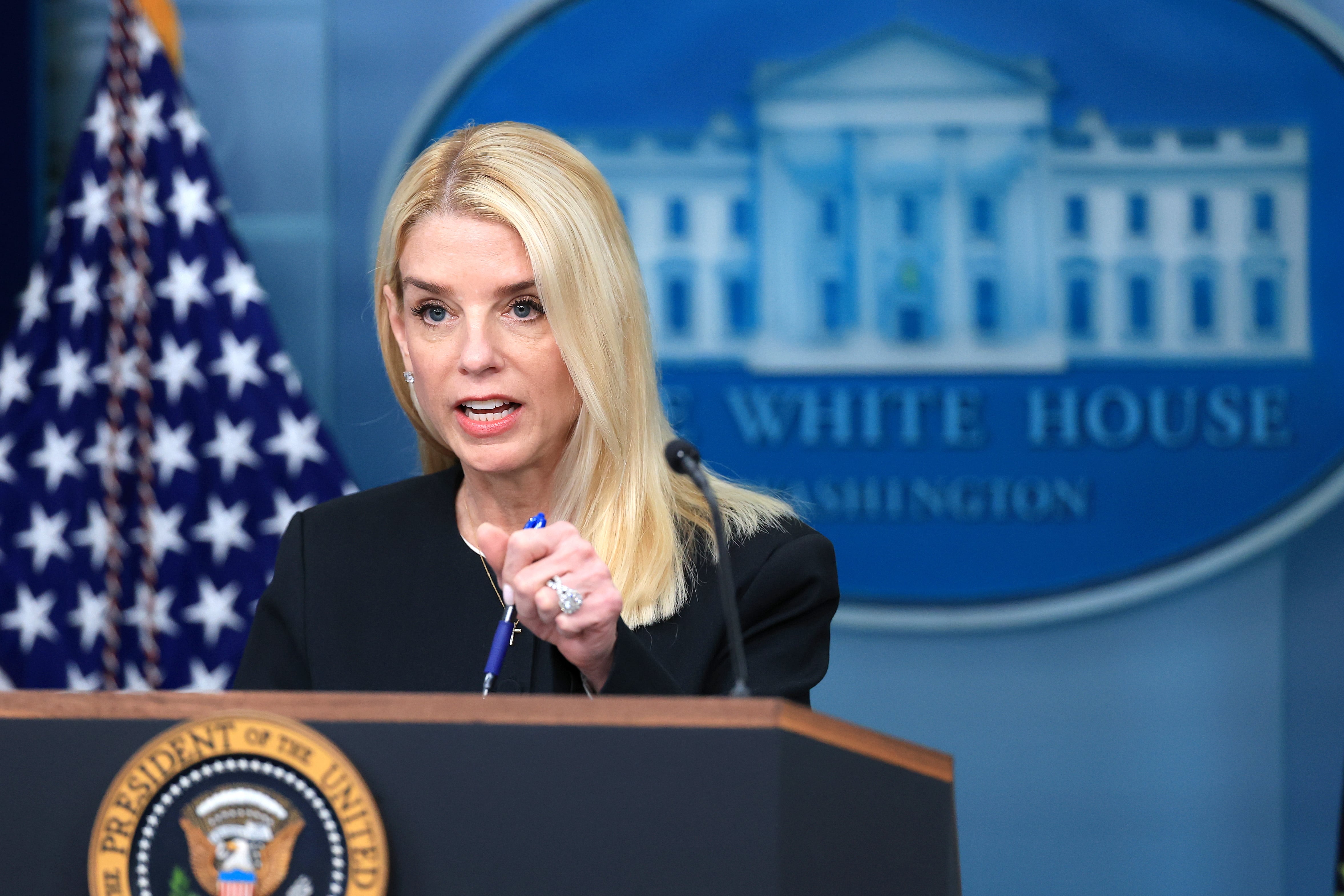 A woman with blonde hair and a dark suit jacket speaks from behind a podium in front of the White House symbol.