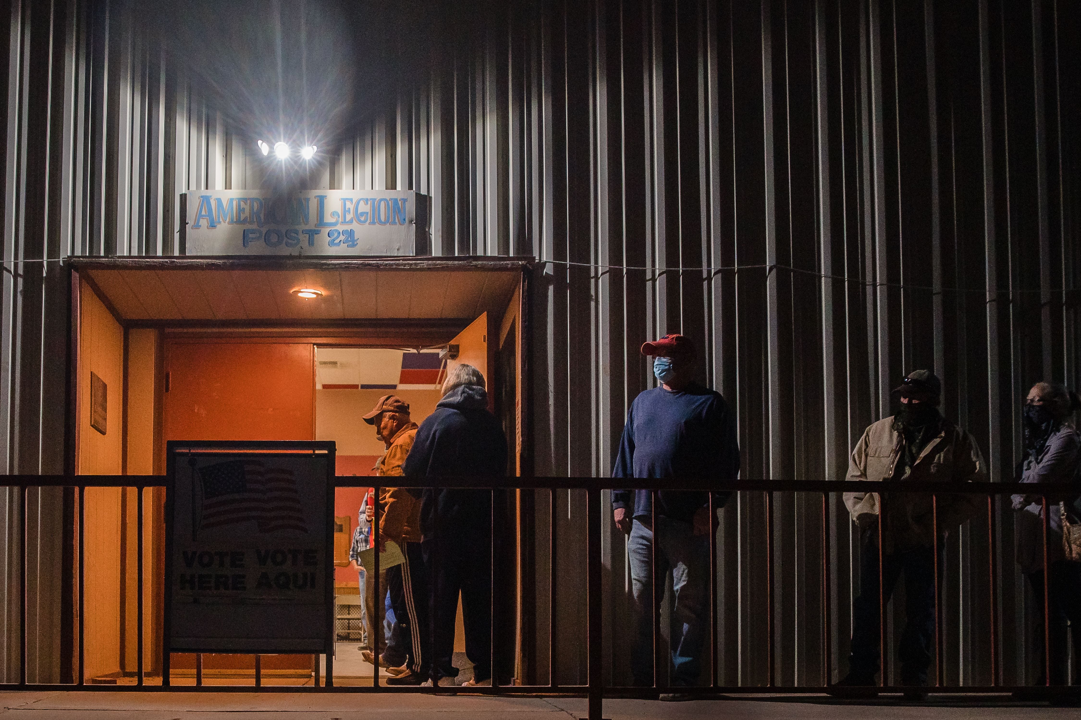 Line of people in warm clothing wait outside building in the dark.